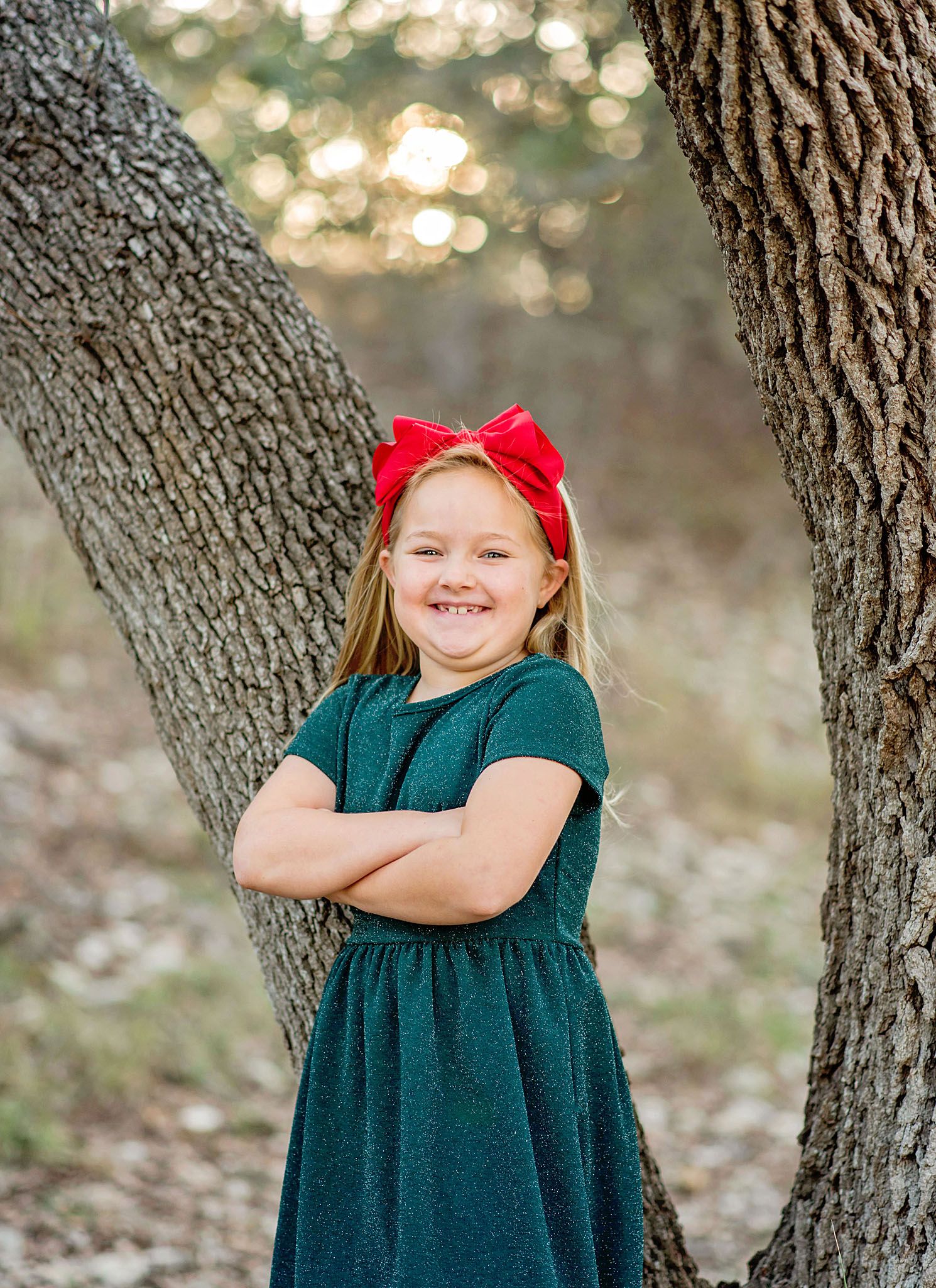 A child in a dark green dress poses playfully against a tree trunk in a sunlit outdoor setting.