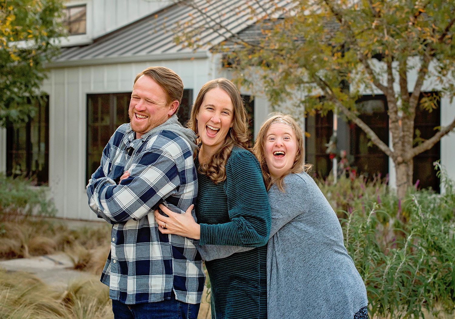 Three people laughing and hugging outdoors in front of a white farmhouse surrounded by autumn trees.