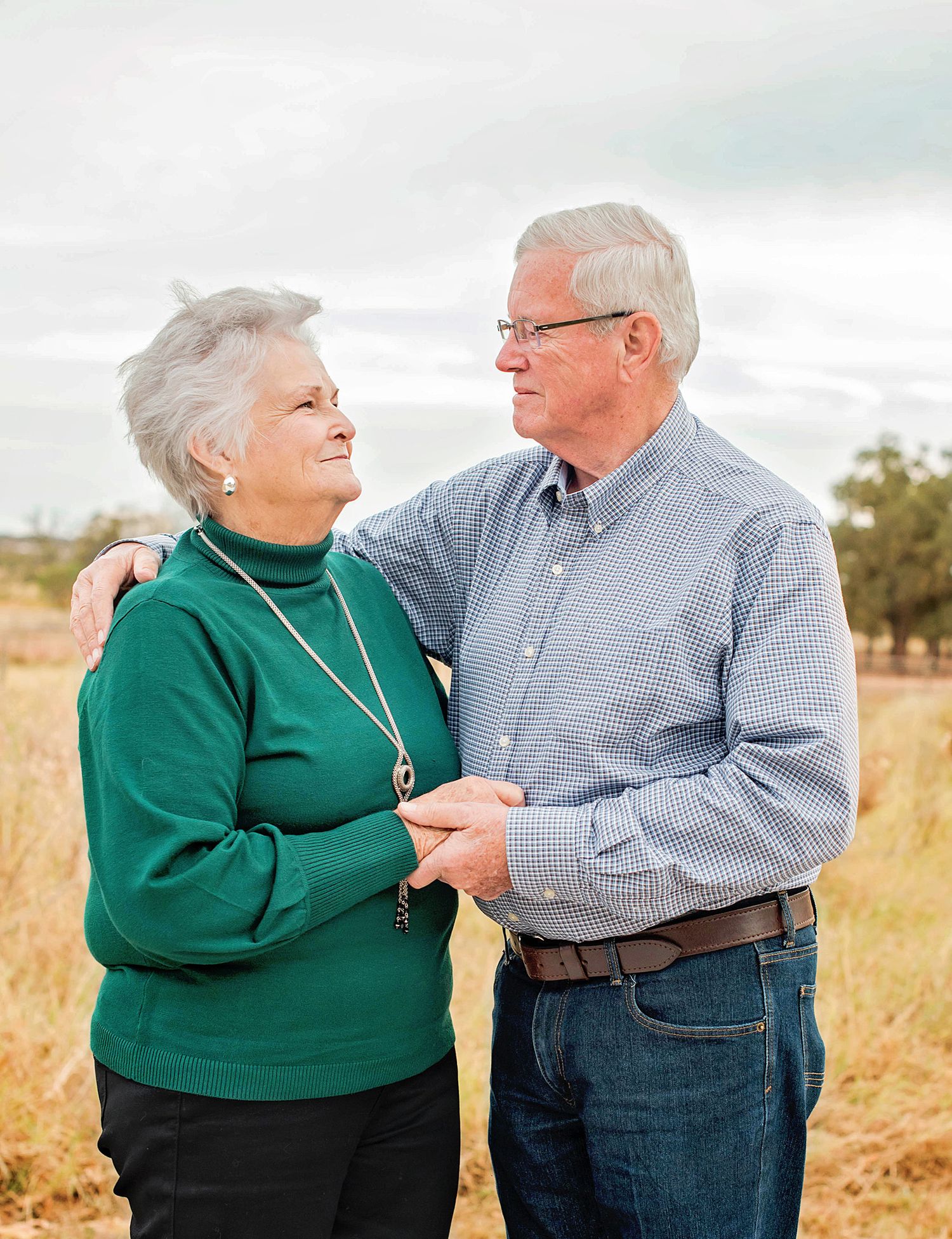 Elderly couple sharing a tender moment in a golden field, smiling at each other with arms around one another.