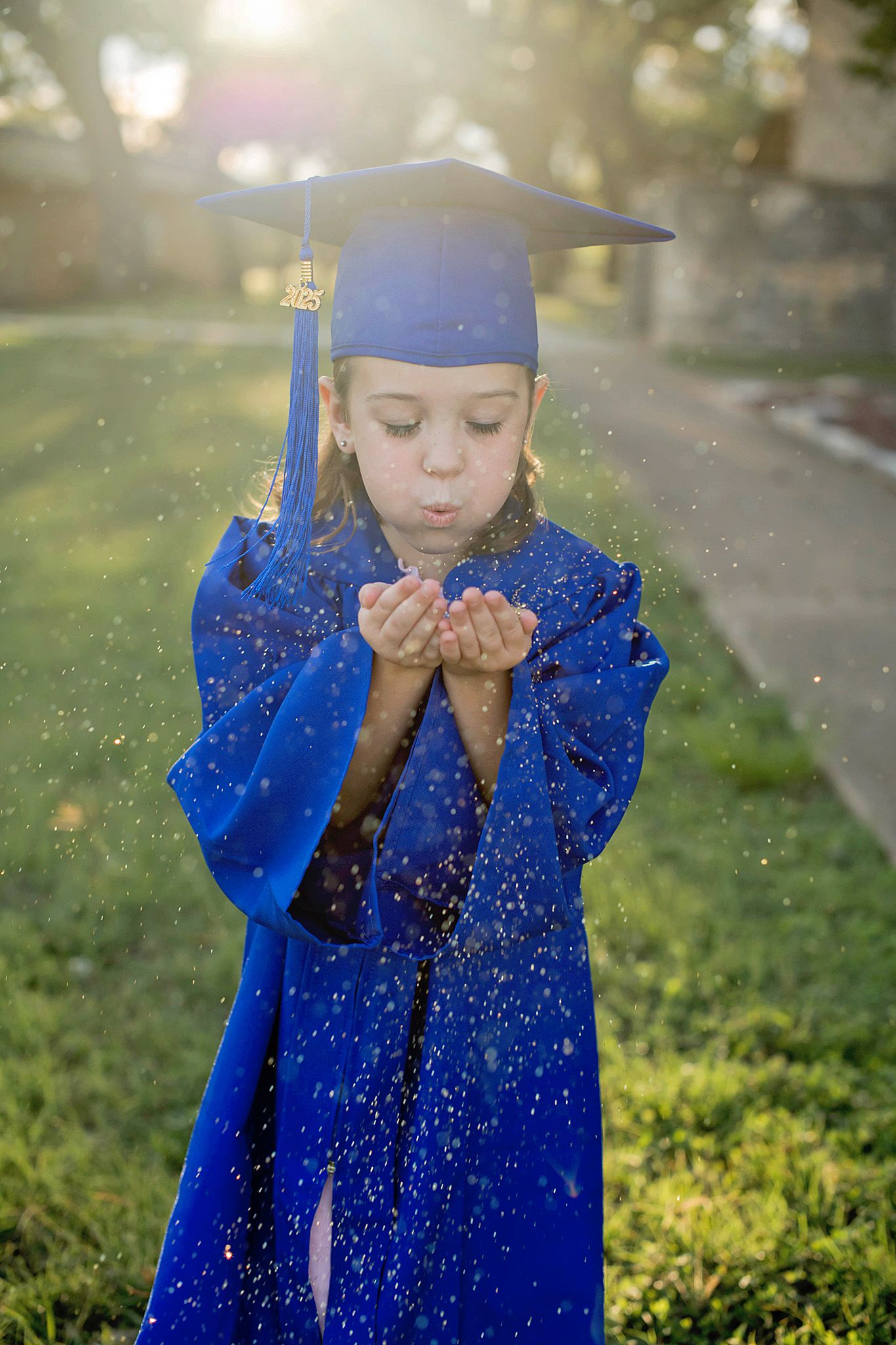 Graduate in royal blue cap and gown blowing glitter in sunlit garden creating magical sparkles in the air.