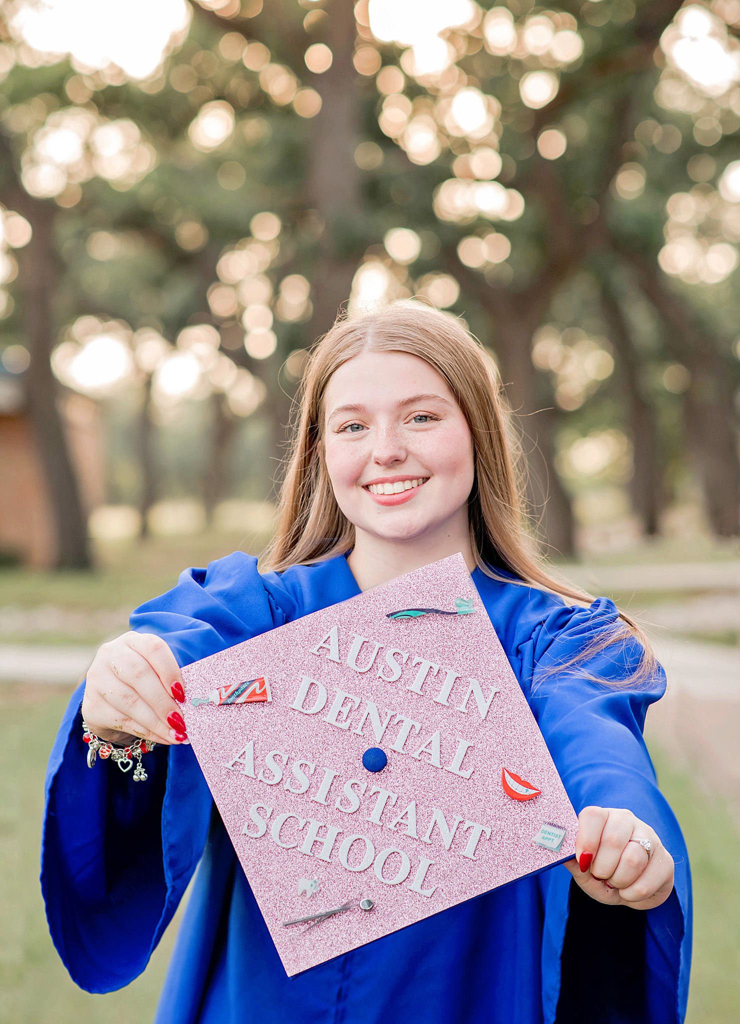 Young woman in blue graduation gown holds decorated cap reading Austin Dental Assistant School outdoors.