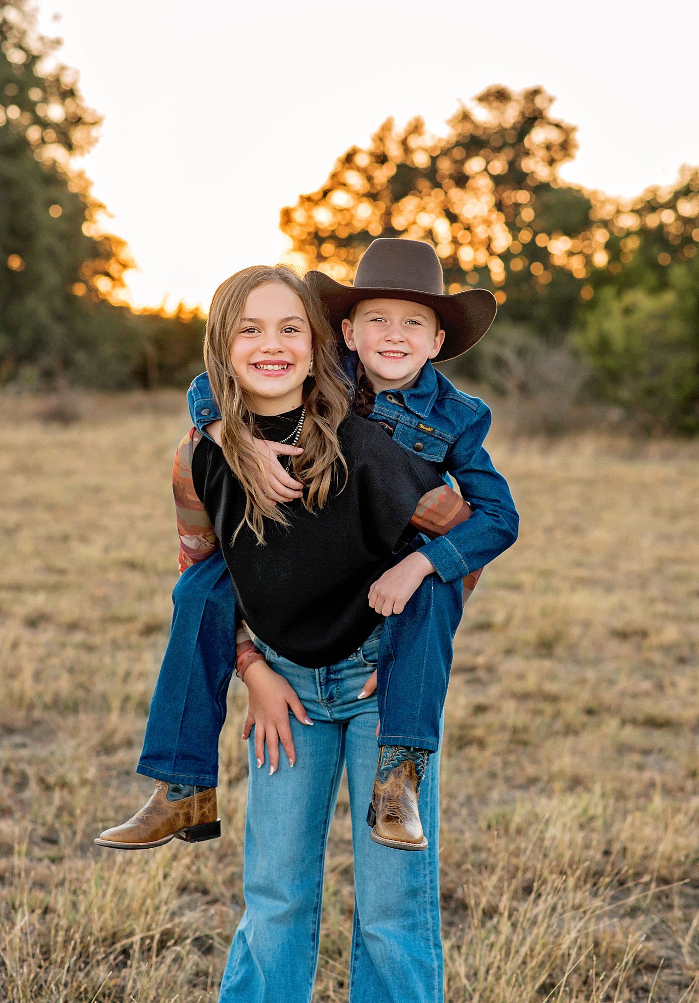 Two children in western wear share playful moments in a golden field at sunset in Hye, TX.
