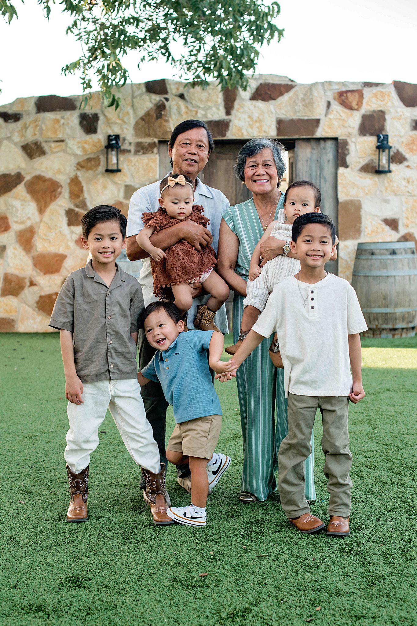Grandparents smiling outdoors with five grandchildren on green grass near a stone wall.