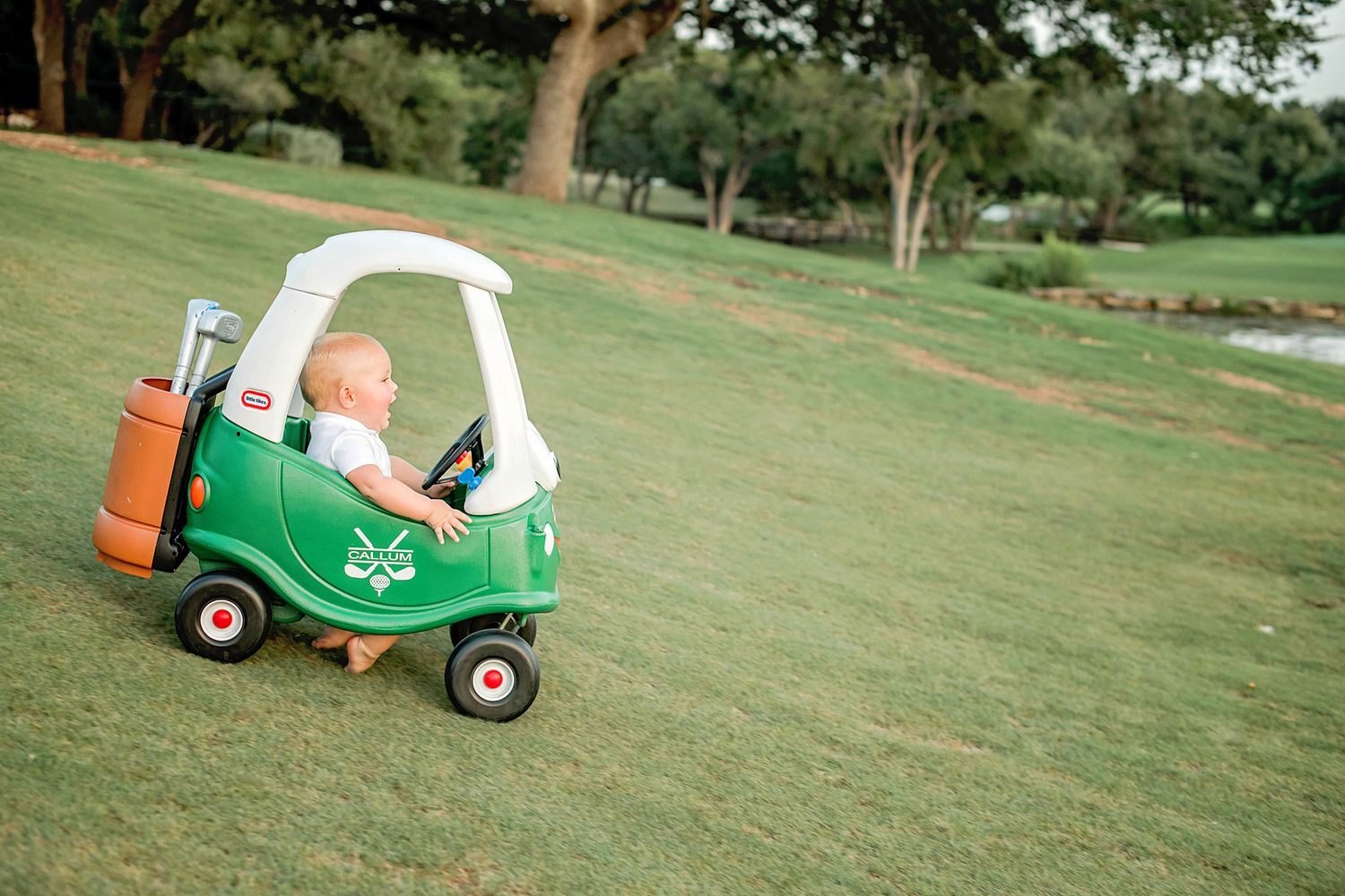 A small child rides in a green toy cozy coupe car on a grassy lawn during a sunny day.