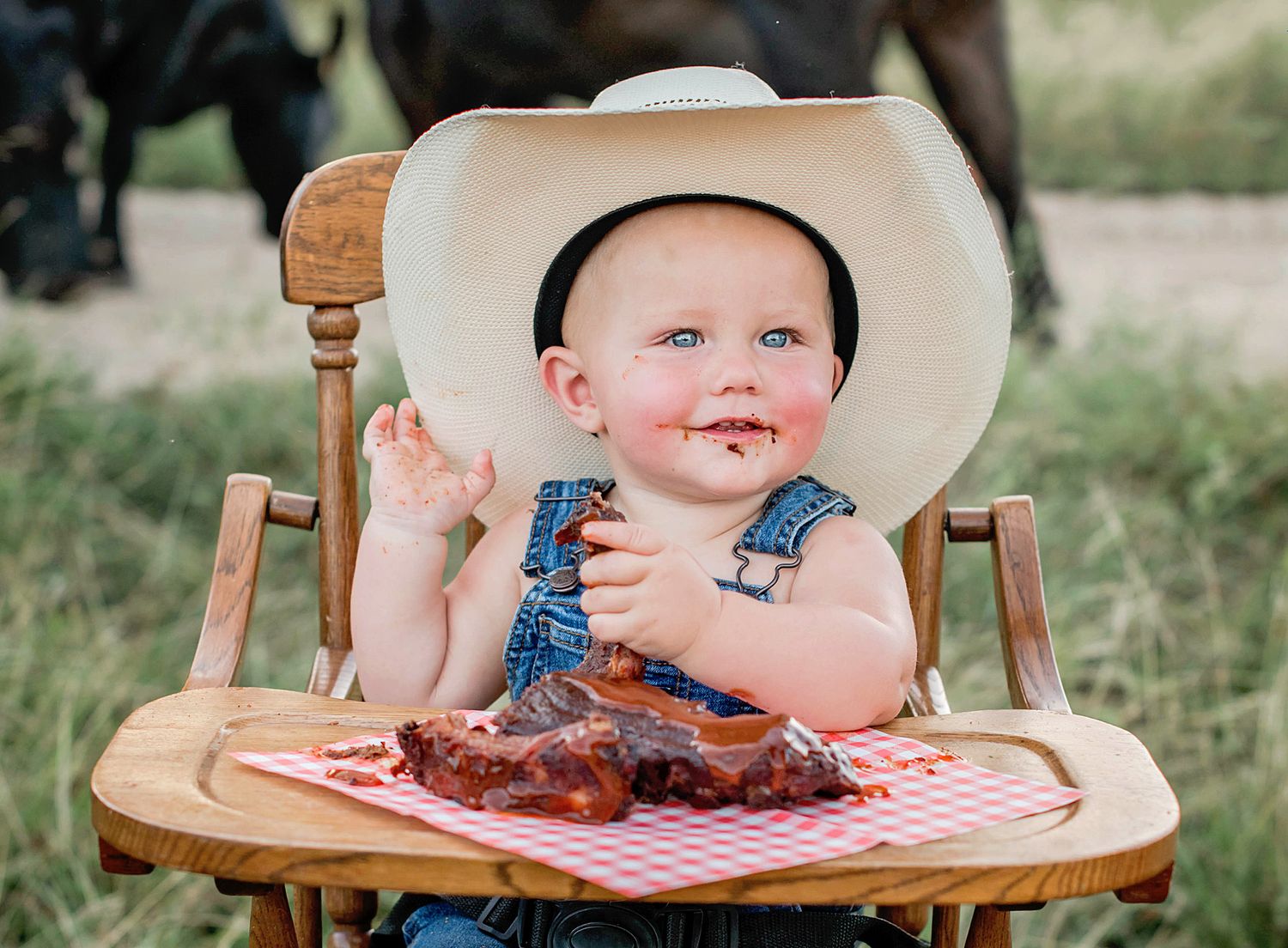 Small child seated in wooden high chair with red and white checkered surface enjoying messy bbq ribs.