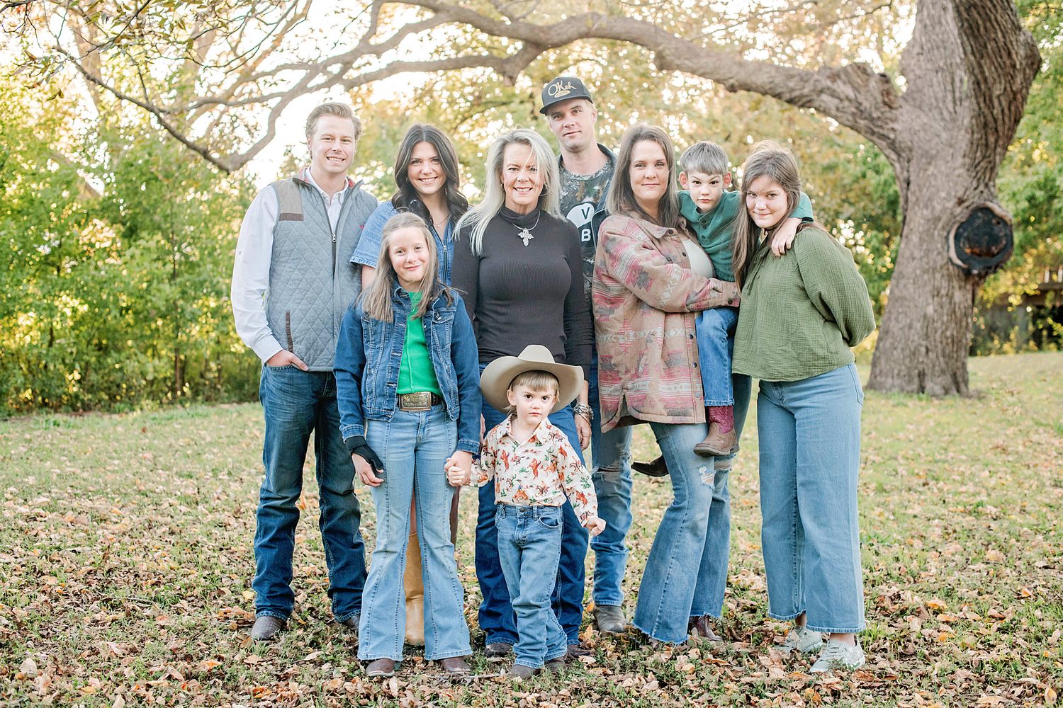 Family of eight posing outdoors in casual western attire among autumn trees.