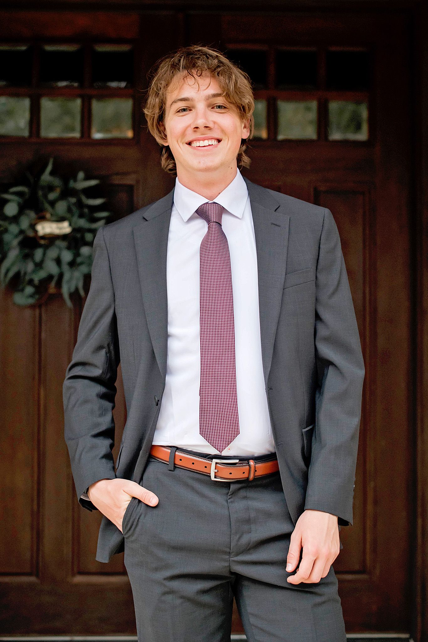 Young man in gray suit with burgundy tie smiling confidently in front of wooden doors.