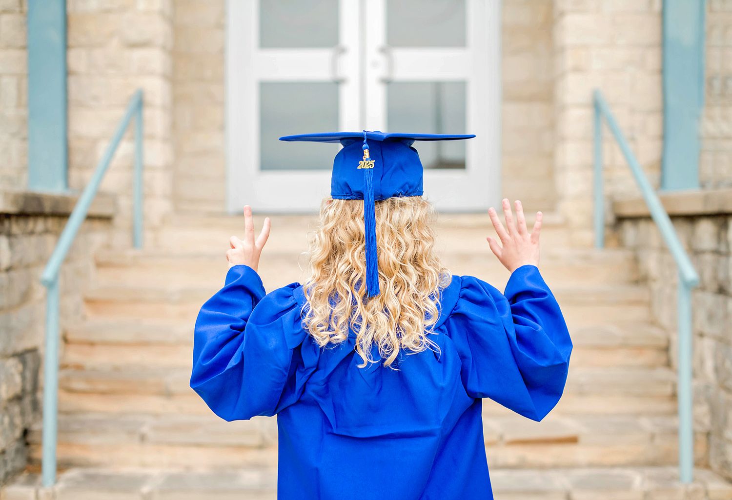 Person in bright royal blue graduation gown raises their arms in celebration against a stone building backdrop.