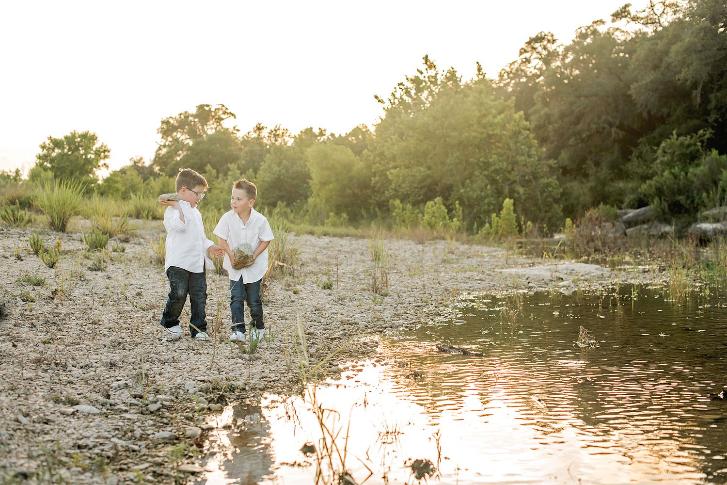 Two boys playing by a rocky riverbank at golden hour with lush green trees in the background.
