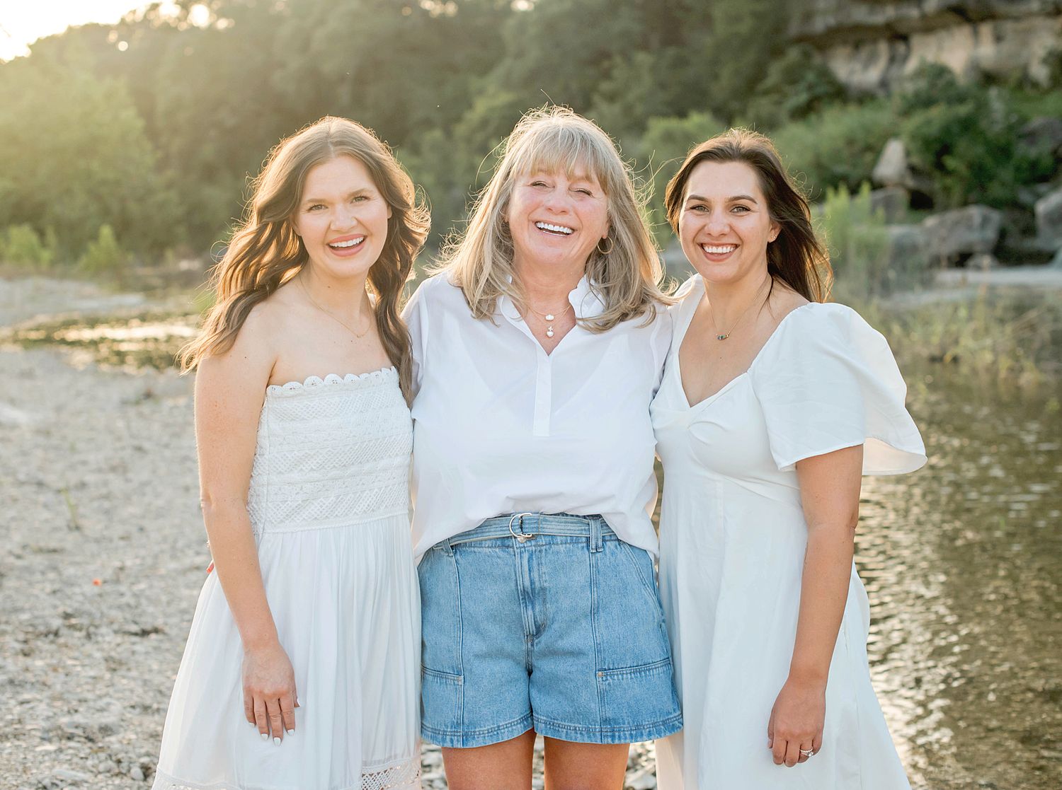Three smiling women in white outfits pose together outdoors near a rocky creek at golden hour.
