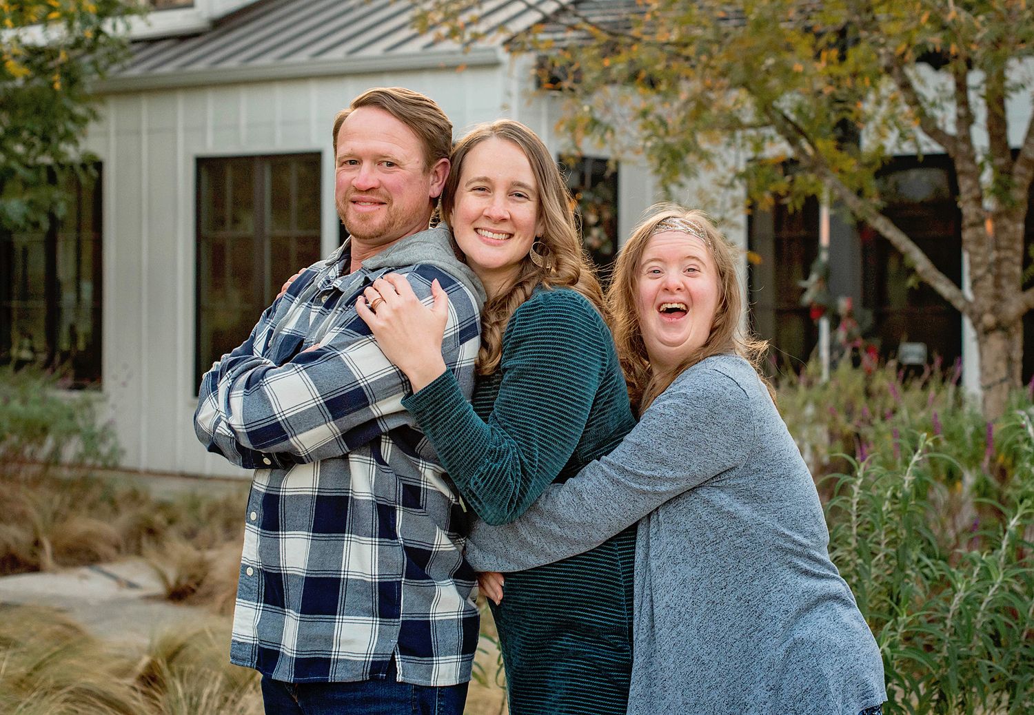 Three smiling people pose together outdoors near a white farmhouse, sharing a warm embrace in a fall setting.