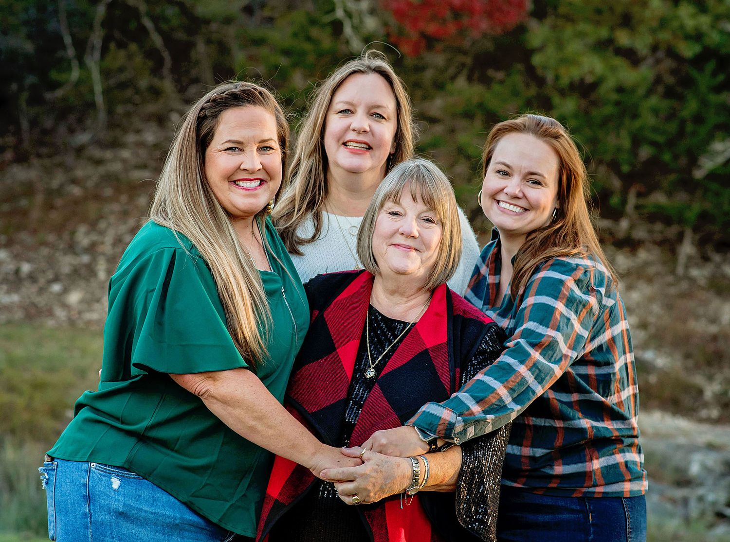 Four women smiling and embracing outdoors in fall setting, wearing casual clothing in green, red, and plaid.