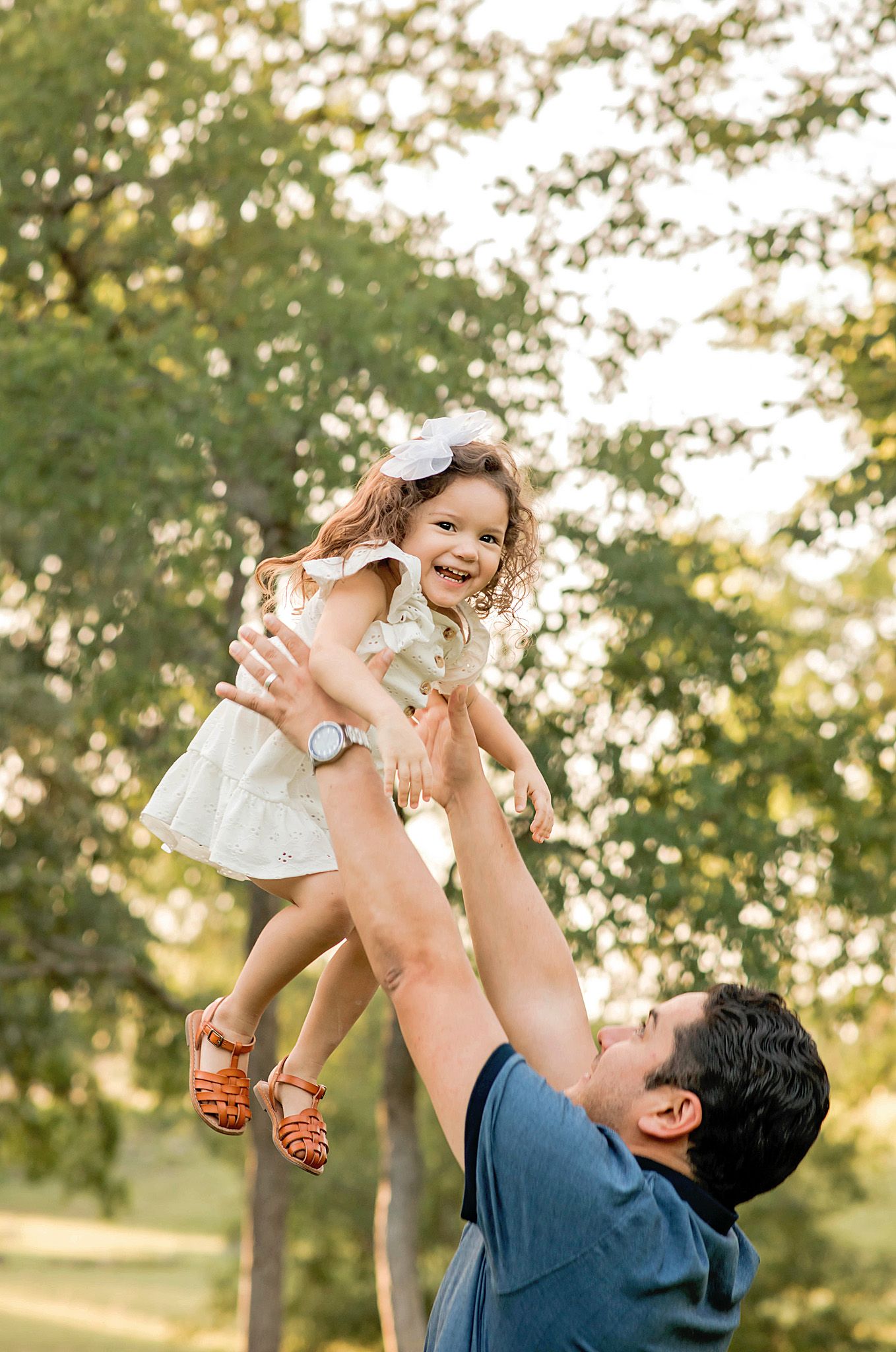 Parent lifts child playfully in the air during golden hour in a park setting with trees in the background.