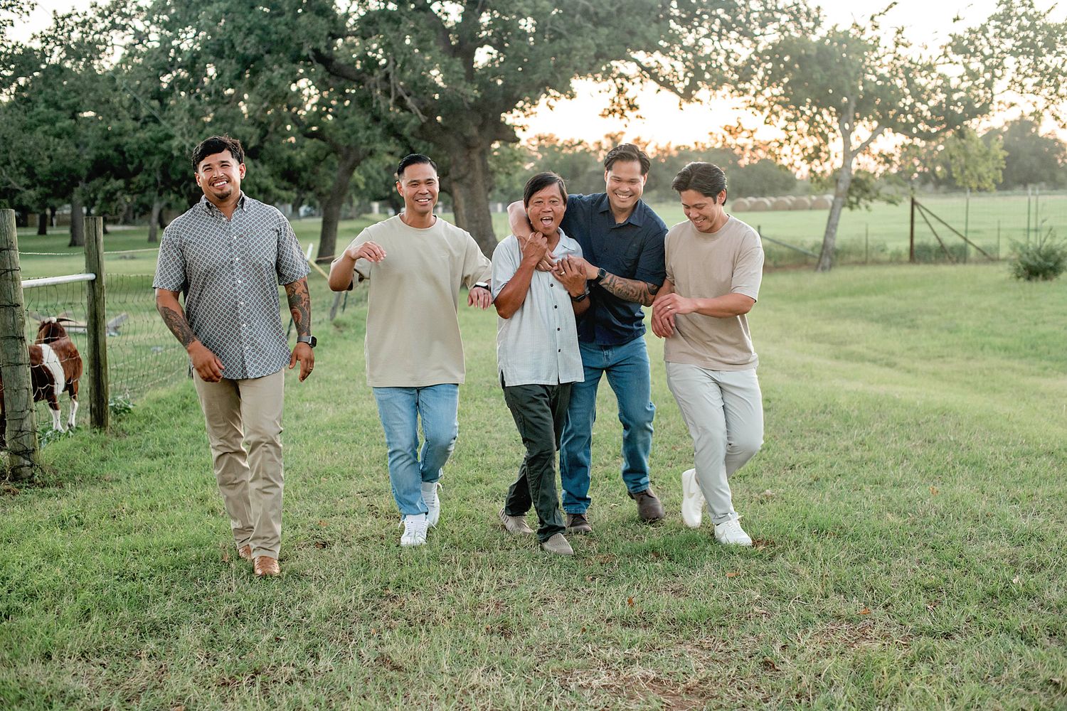 Five men walking and laughing together outdoors on a green lawn at sunset near trees and a fence.