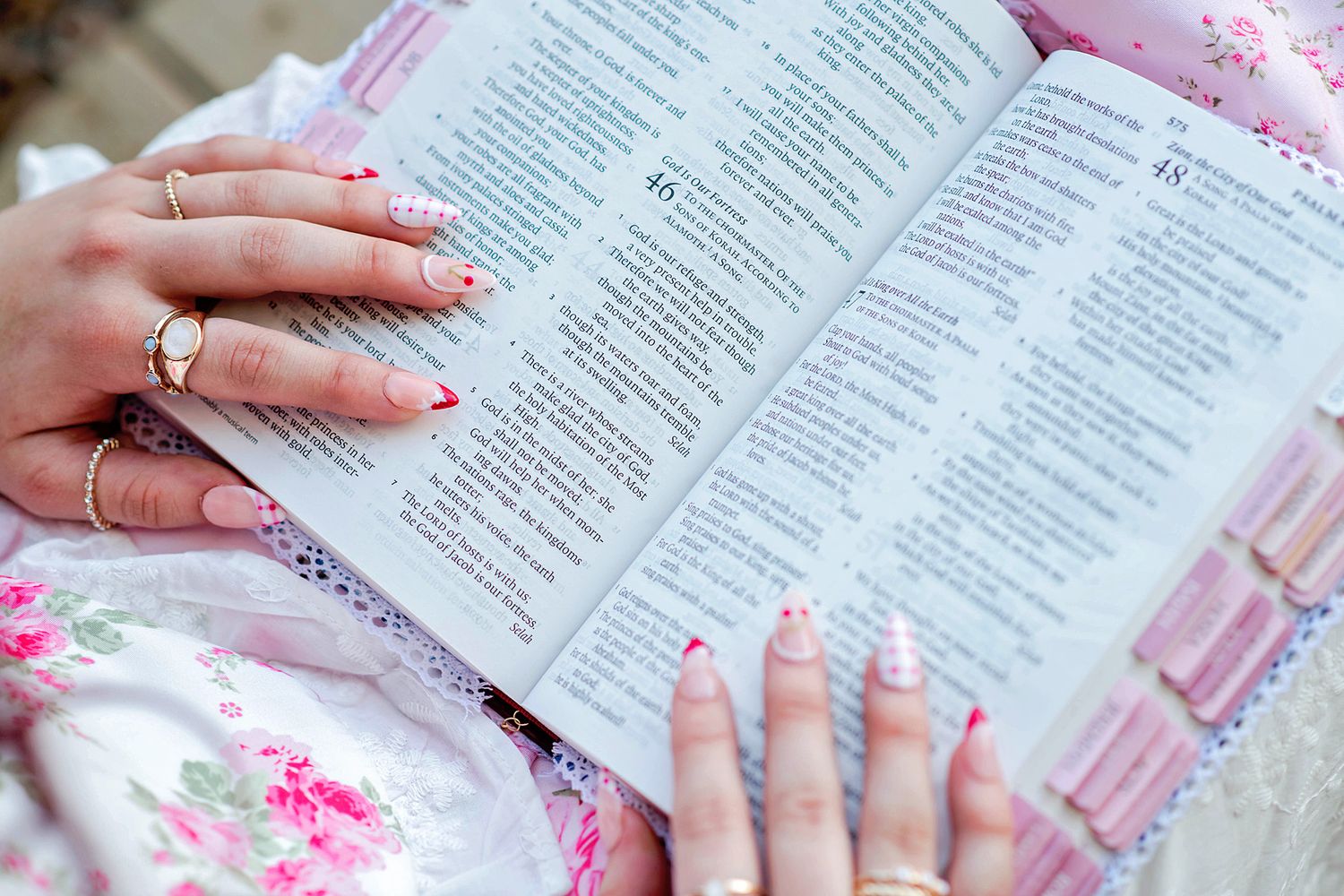 Woman with red manicured nails and rings reading an open Bible while wearing a floral pink dress.