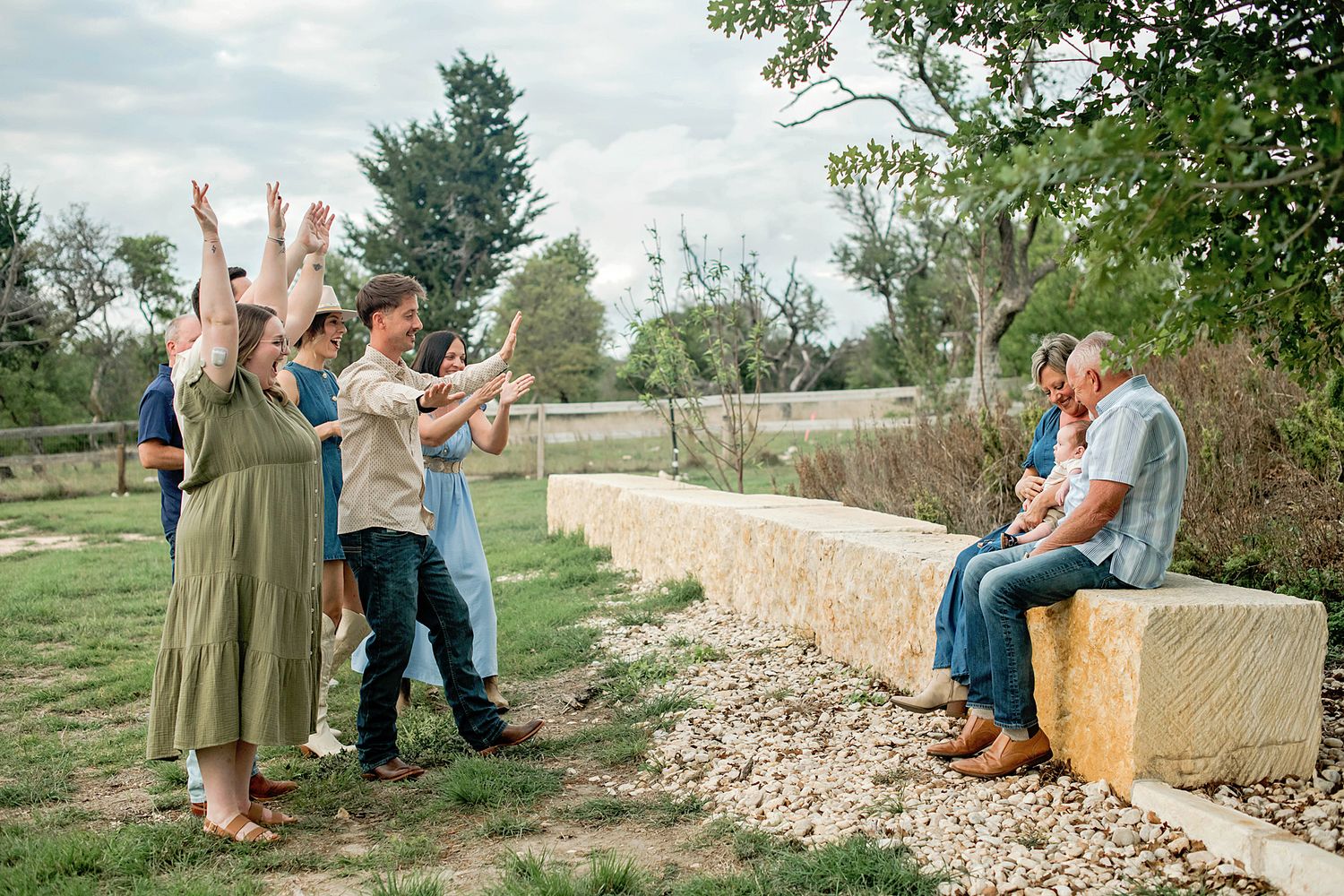 Family group celebrating outdoors on a farm, with people cheering toward an elderly couple seated on a stone bench.