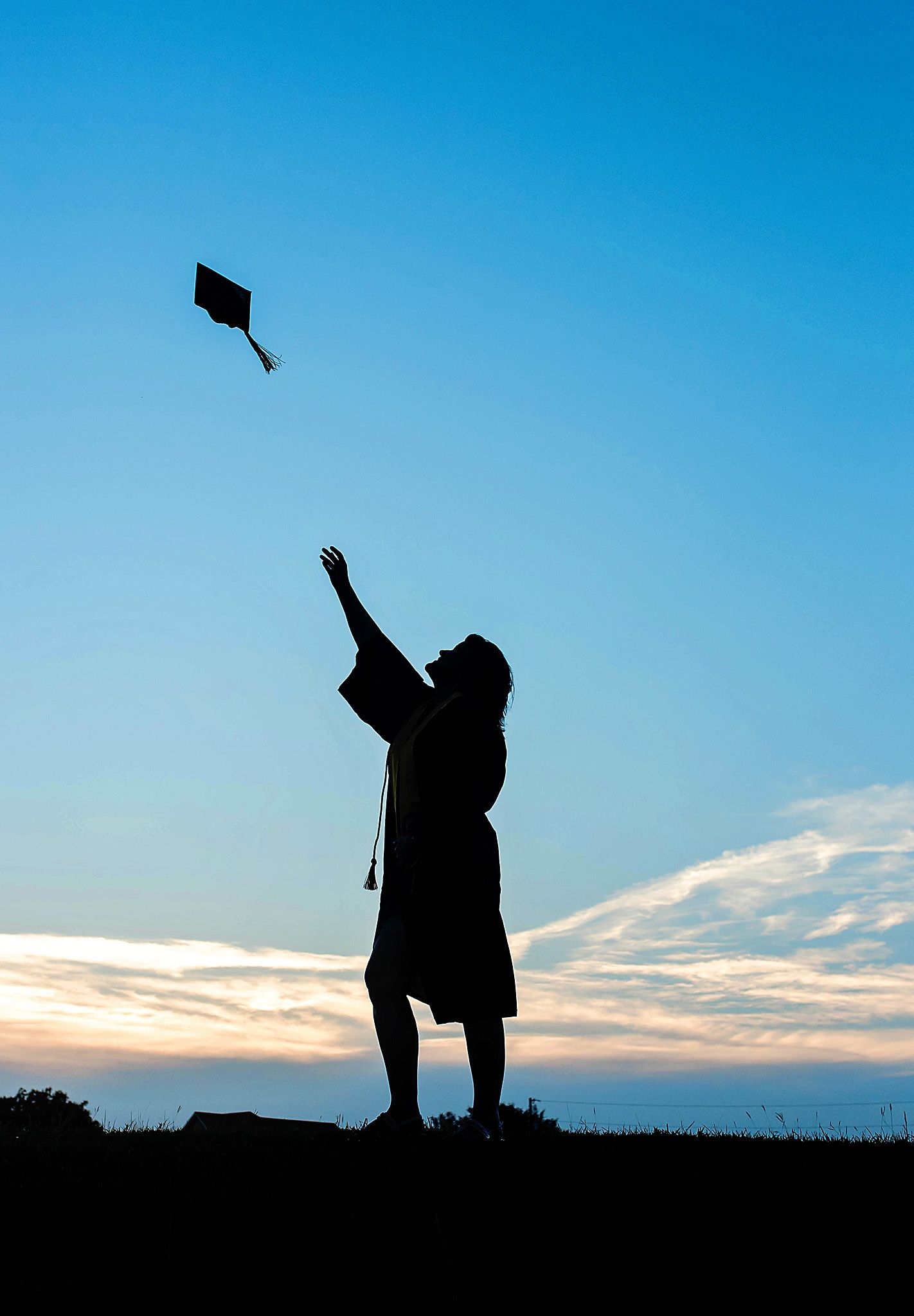Silhouette of a graduate in cap and gown tossing mortarboard into blue sky at sunset.
