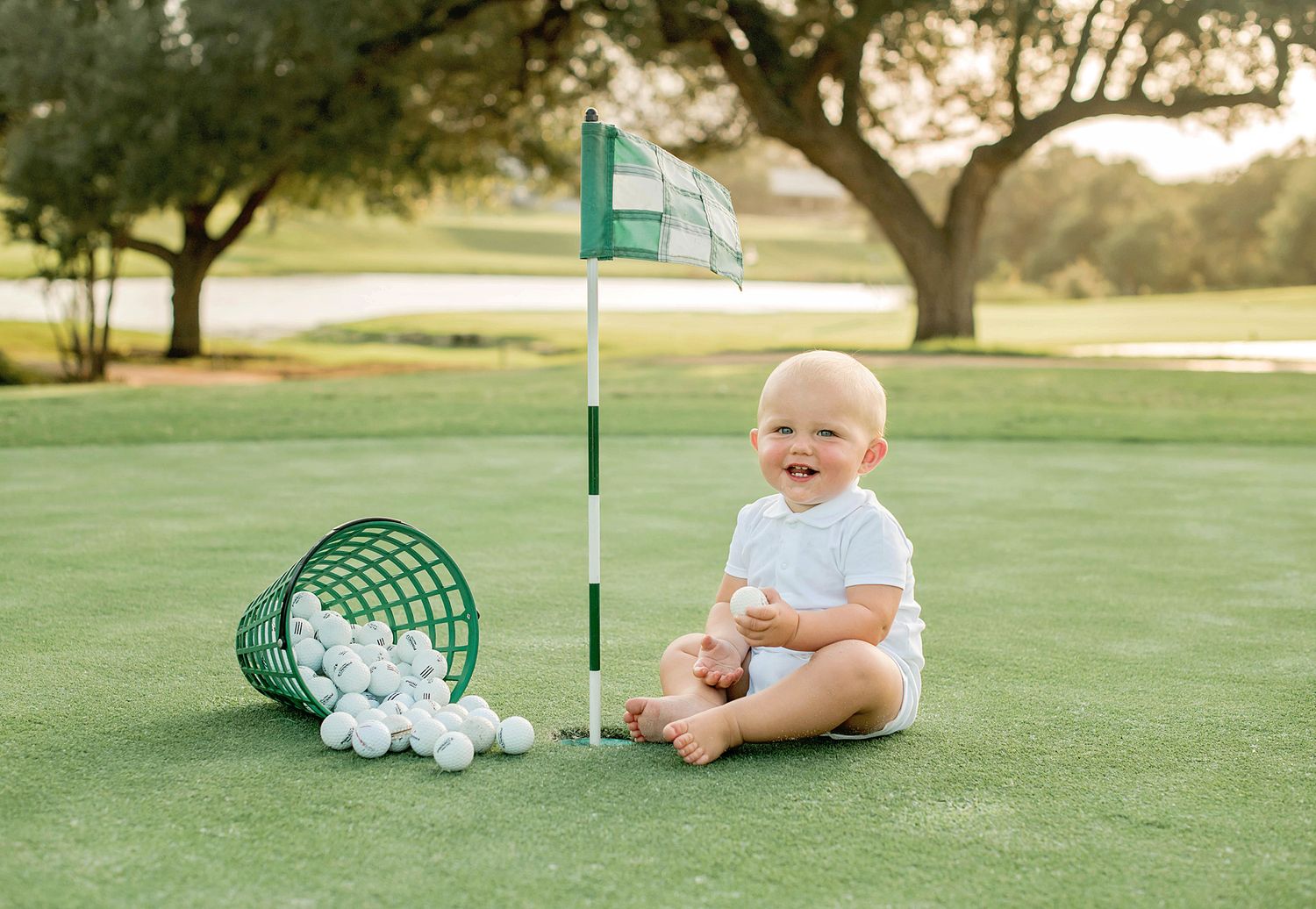 A smiling baby sits on a golf course green next to a golf flag and practice balls during golden hour lighting.