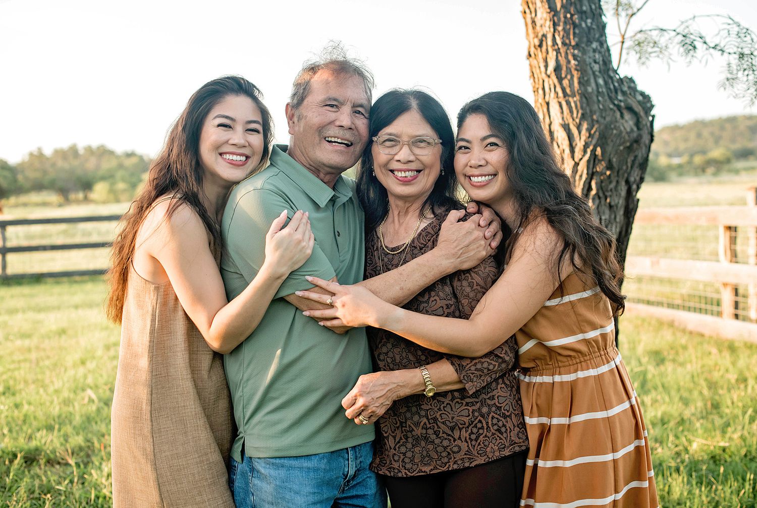 Asian family of four smiling and embracing near a tree in a sunny outdoor field setting.