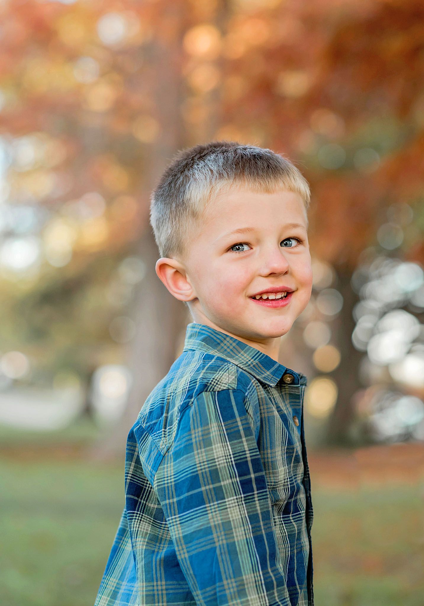 A child wearing a blue plaid shirt smiles while standing outdoors against a warm autumn background.