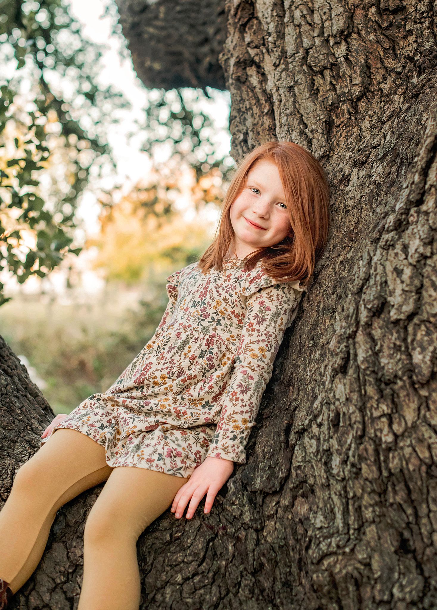 Person in floral top and tan pants relaxes against large tree trunk in outdoor autumn setting.