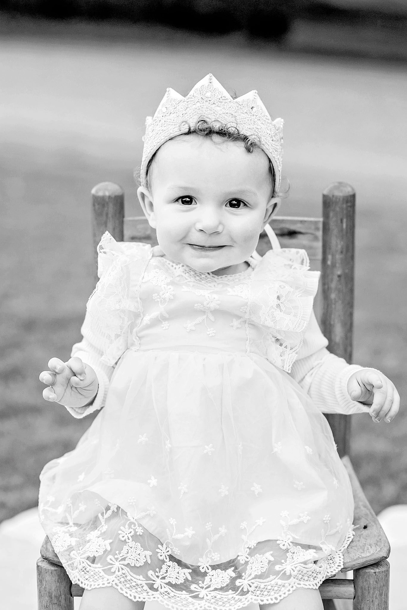 Black and white portrait of a smiling baby girl in a white lace dress and crown sitting on a wooden chair outdoors.