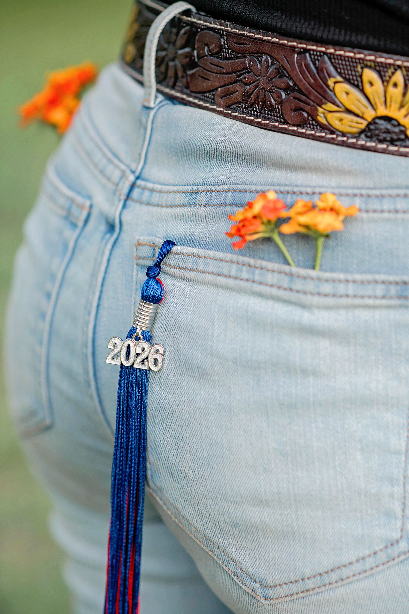 Close-up of light wash jeans with a 2026 graduation tassel charm in the pocket, orange flowers, and a tooled leather belt.