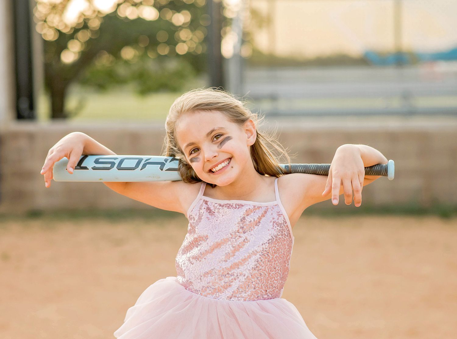Young girl in pink sequin dress holding a baseball bat on her shoulders, smiling outdoors at a softball field.