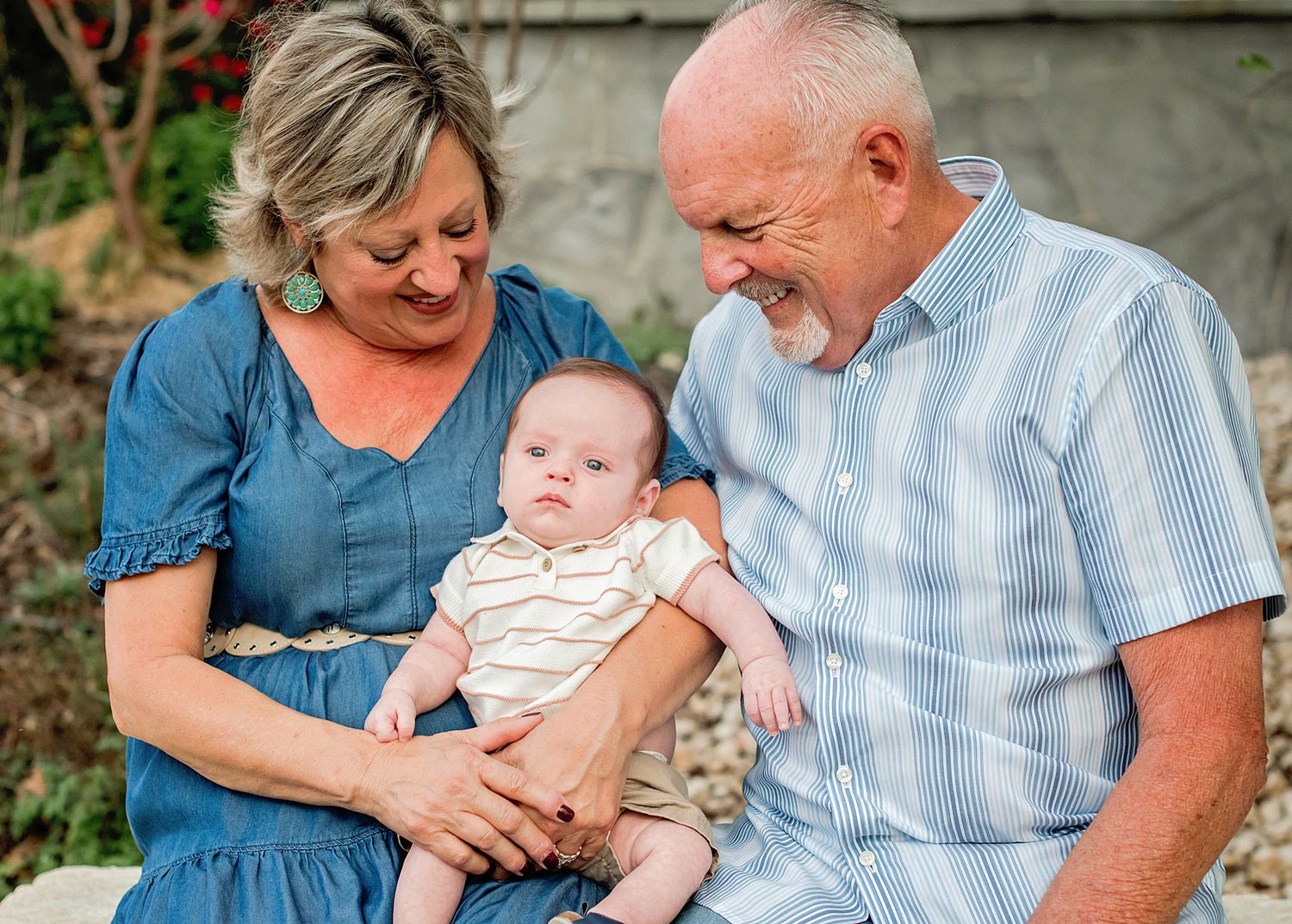 Grandparents smiling and holding a newborn baby outdoors in a garden setting.