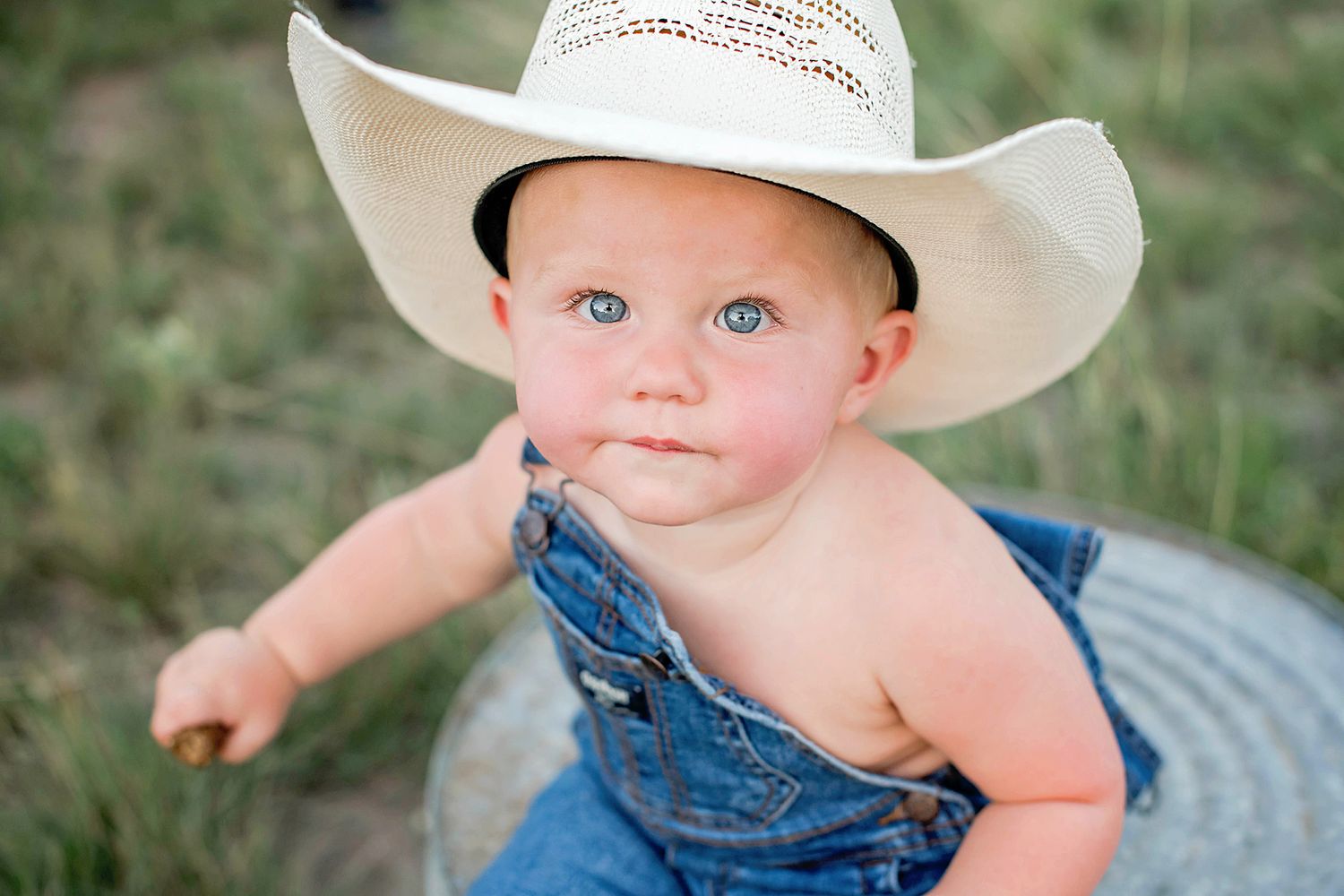 Adorable baby wearing a white cowboy hat and denim overalls sits on grass during outdoor photoshoot.