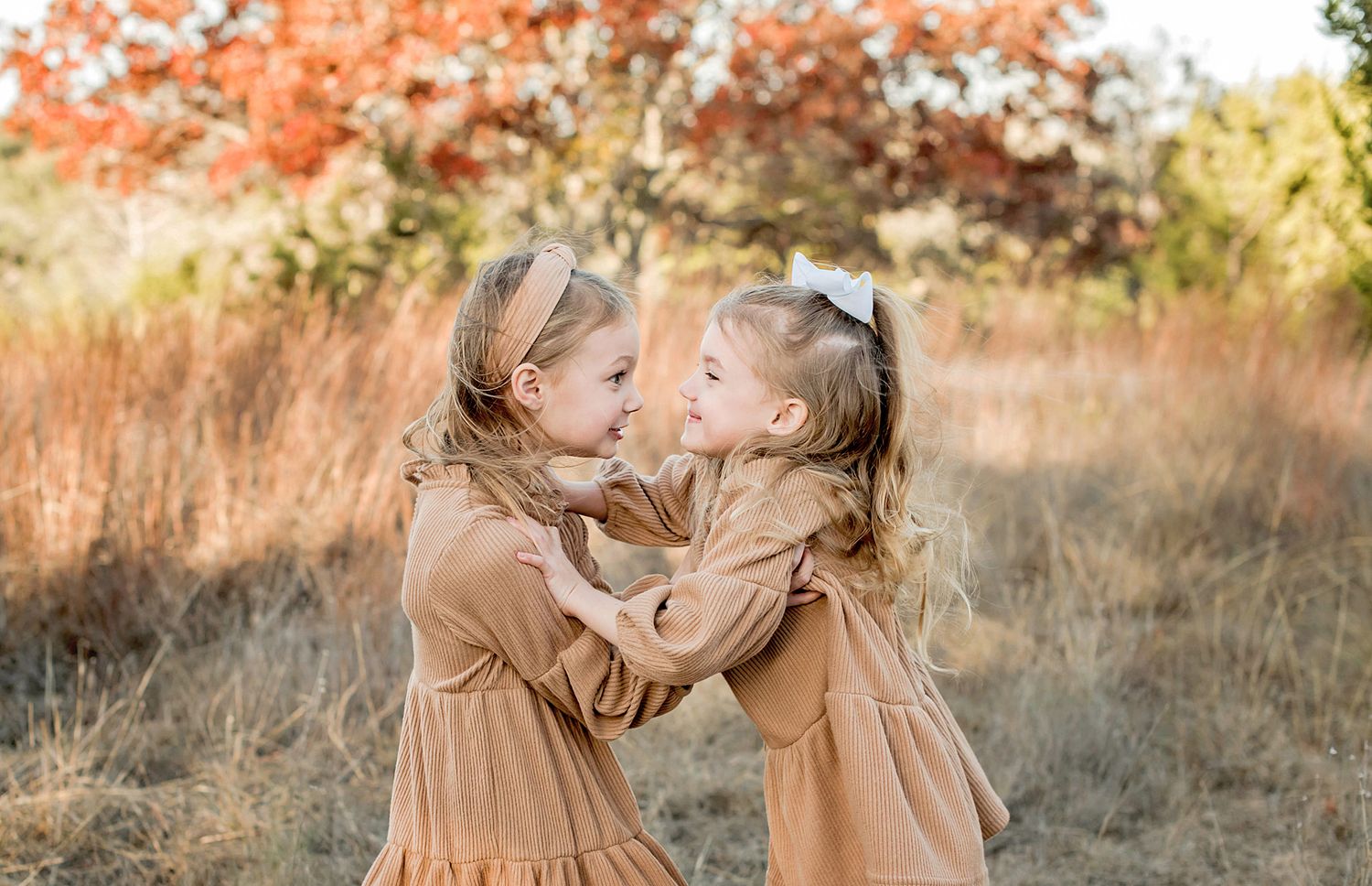 Two children in matching beige dresses play and twirl in a golden field at sunset.
