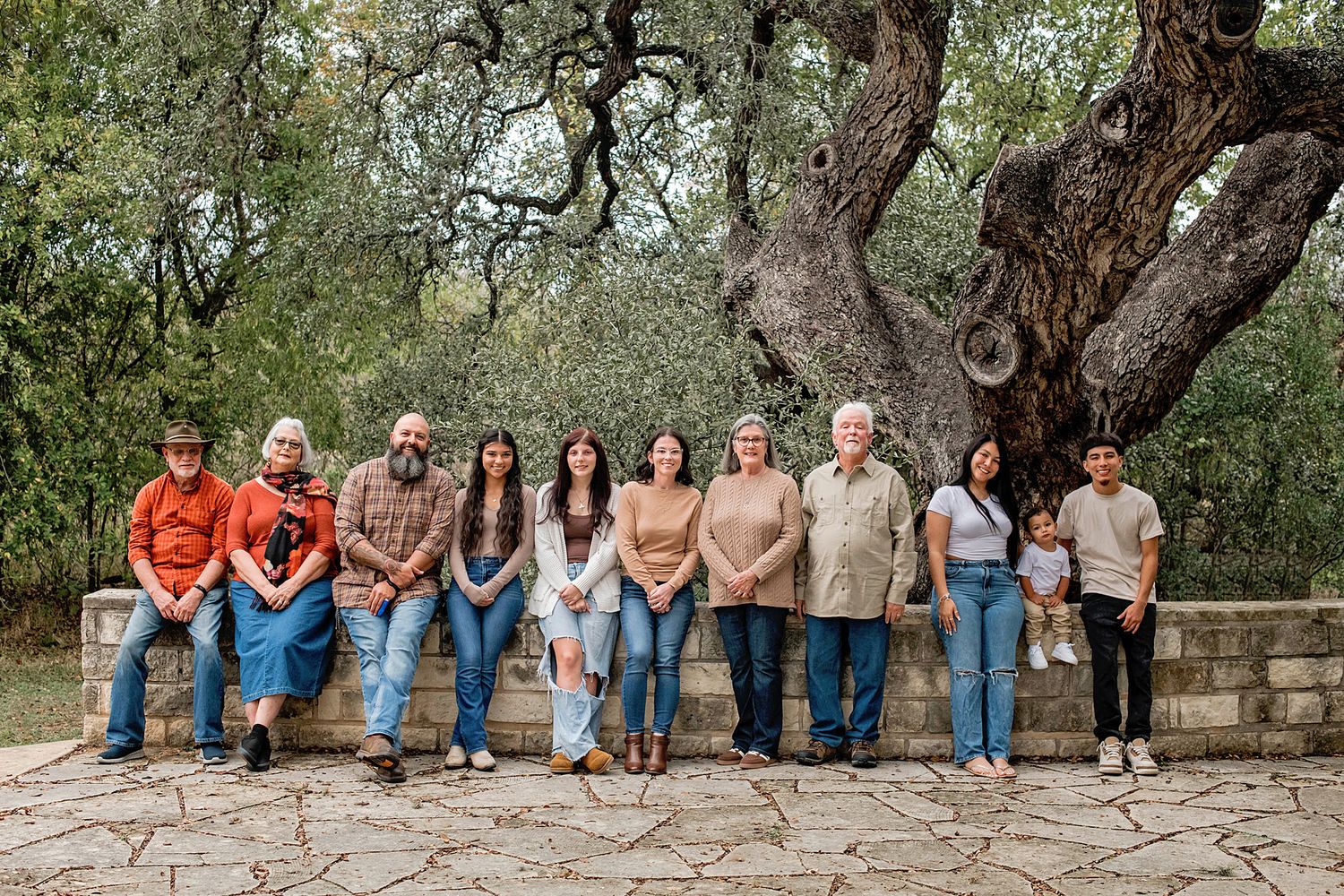 Large family group portrait outdoors on stone patio beneath sprawling oak trees in a natural park setting.
