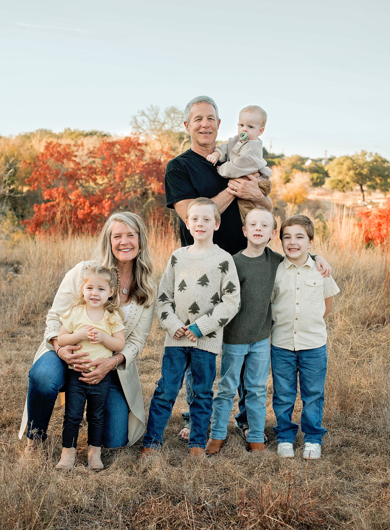 Family of seven posing outdoors in a golden field with autumn foliage, smiling together on a sunny day.