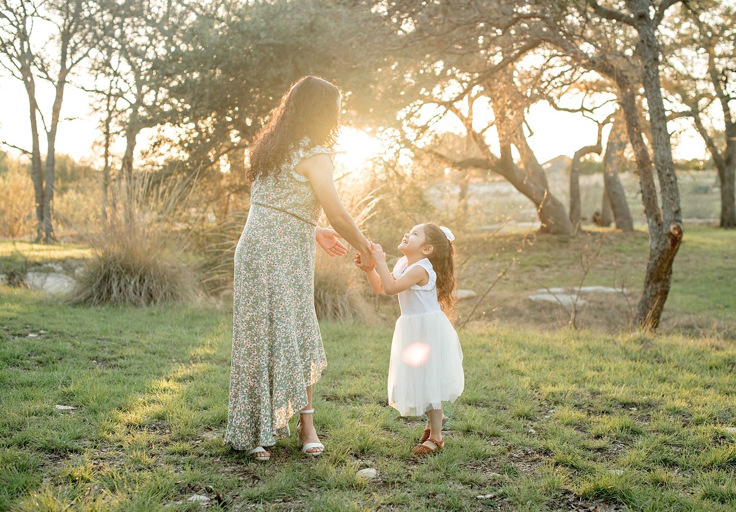 Mother and daughter dancing in golden sunset light in a meadow, girl in white dress holding mom's hands outdoors.