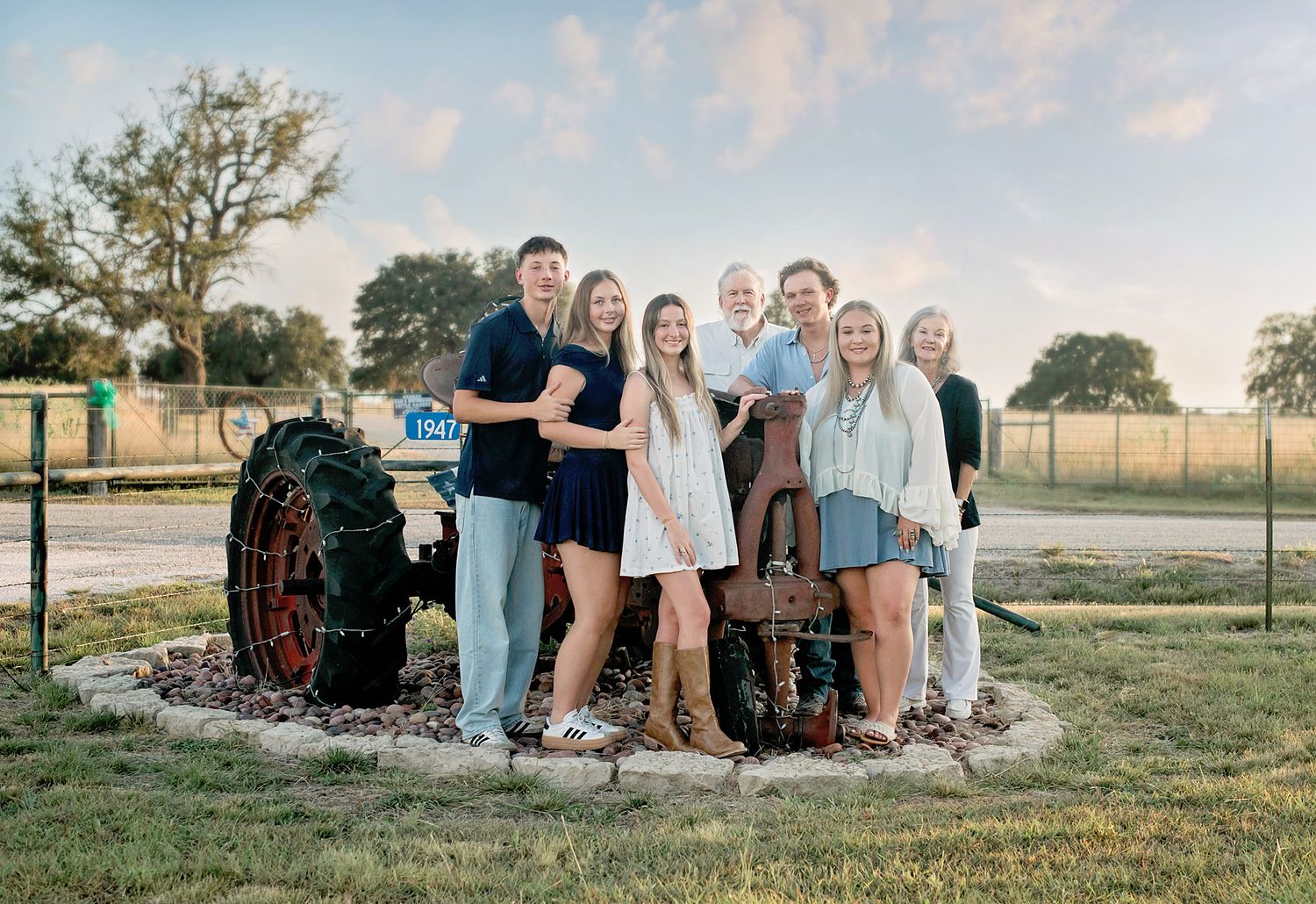 A large family poses together outdoors near a vintage tractor on a green lawn at golden hour.