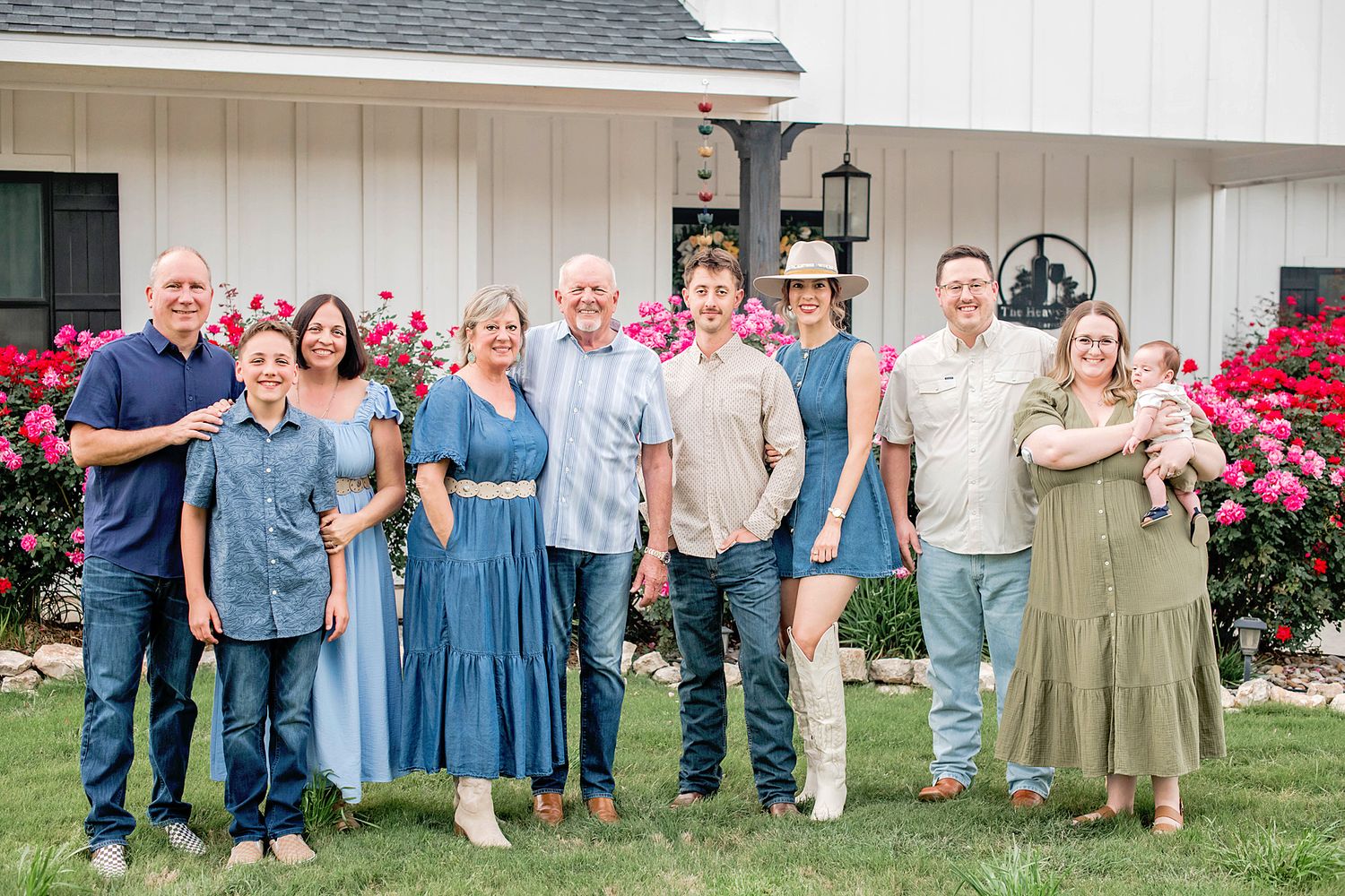 Family of nine posing outdoors in front of a white barn surrounded by blooming pink roses on a sunny day.