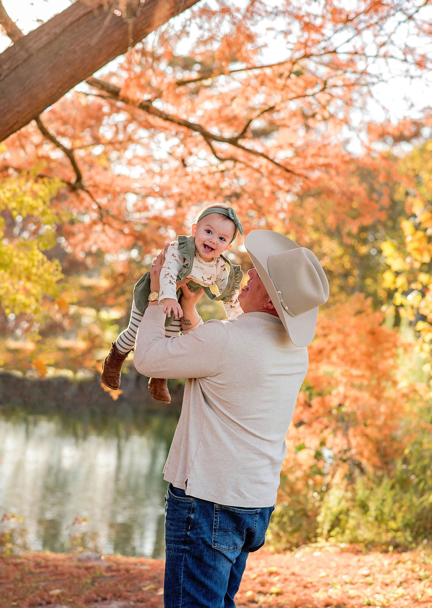 A parent and baby share a tender moment among vibrant orange autumn foliage by a tranquil lake.