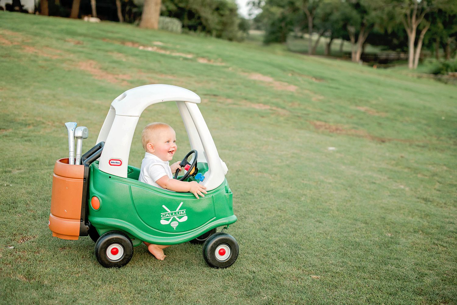 A little green toy Cozy Coupe car sits on a grassy lawn with golf clubs attached to the back.