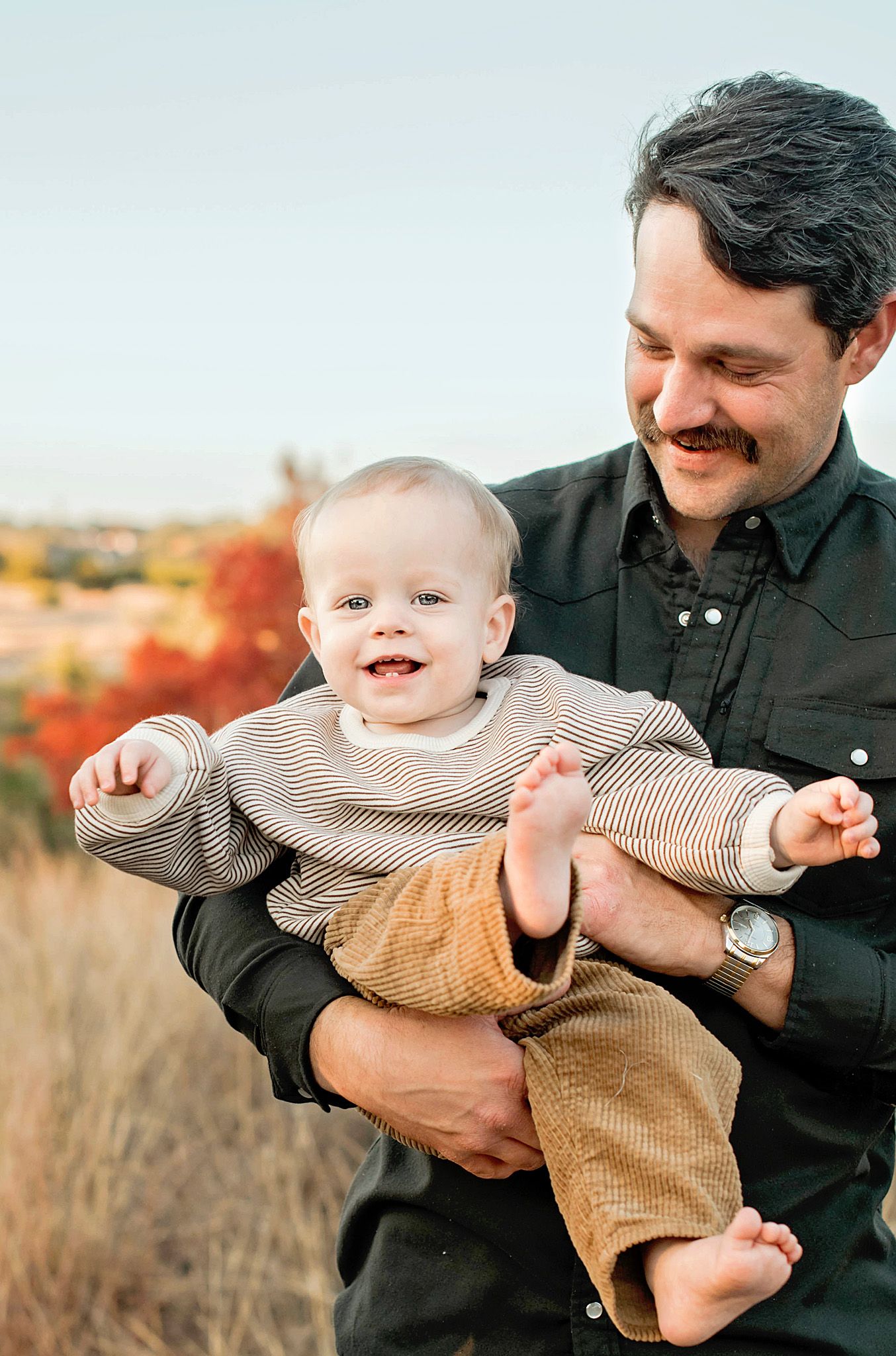 A parent in black clothing playfully holds and lifts a smiling baby wearing a beige sweater in an outdoor autumn setting.