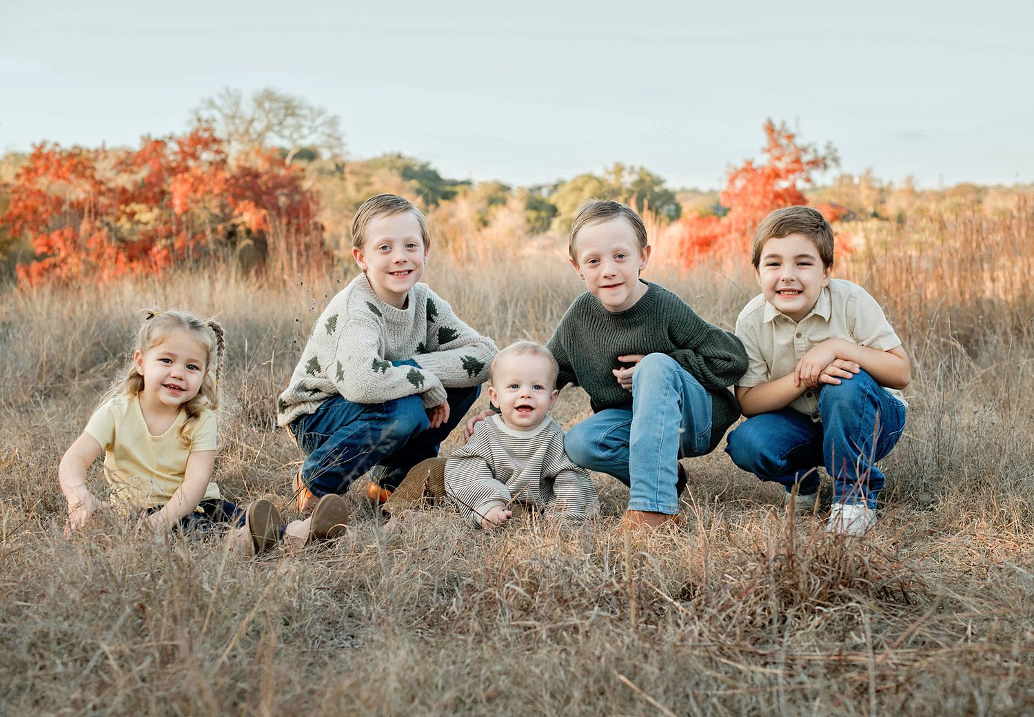 Five children smiling together in a golden autumn field with colorful fall foliage in the background.