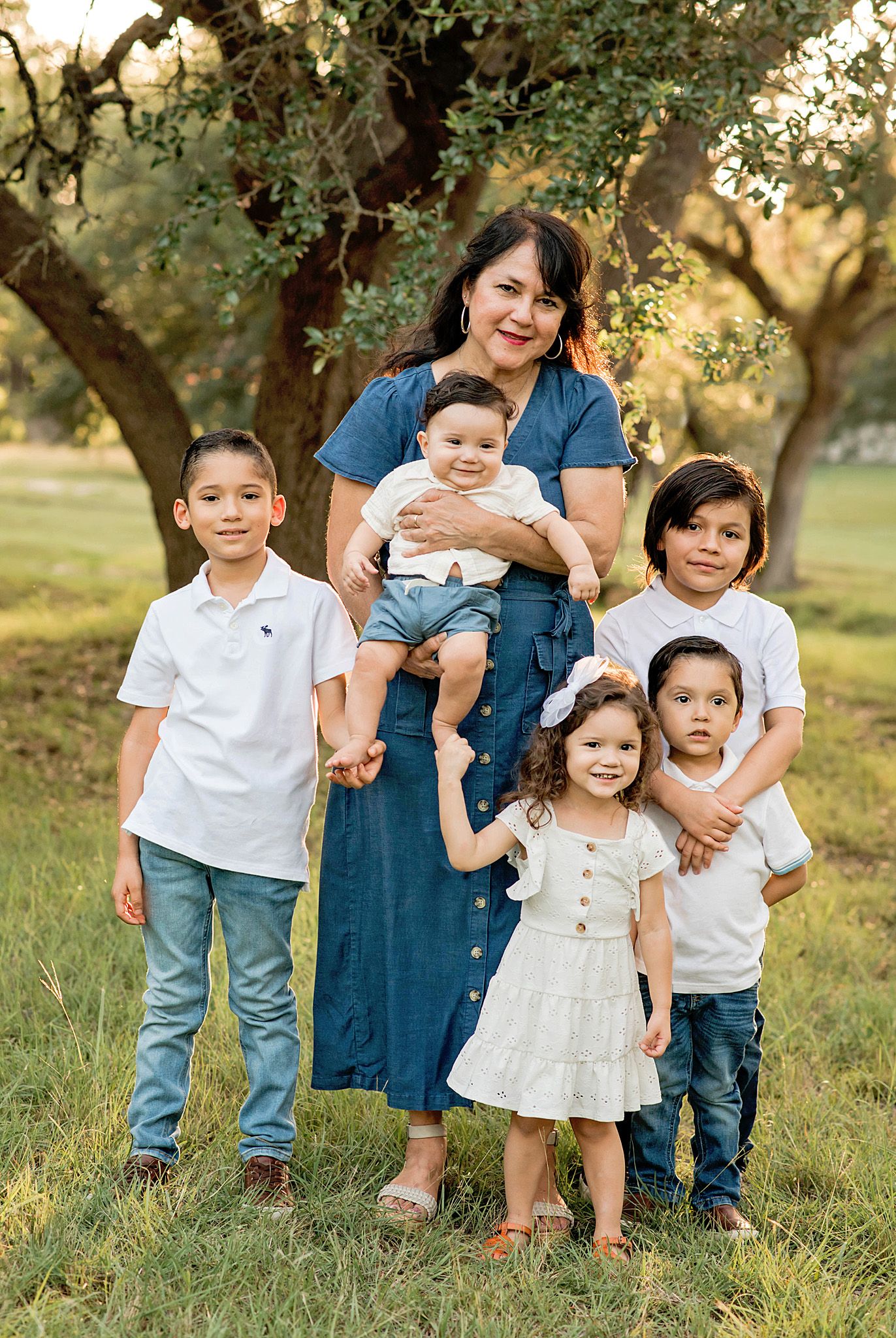 A grandmother poses outdoors with five grandchildren under oak trees in a warm, golden-light family portrait.