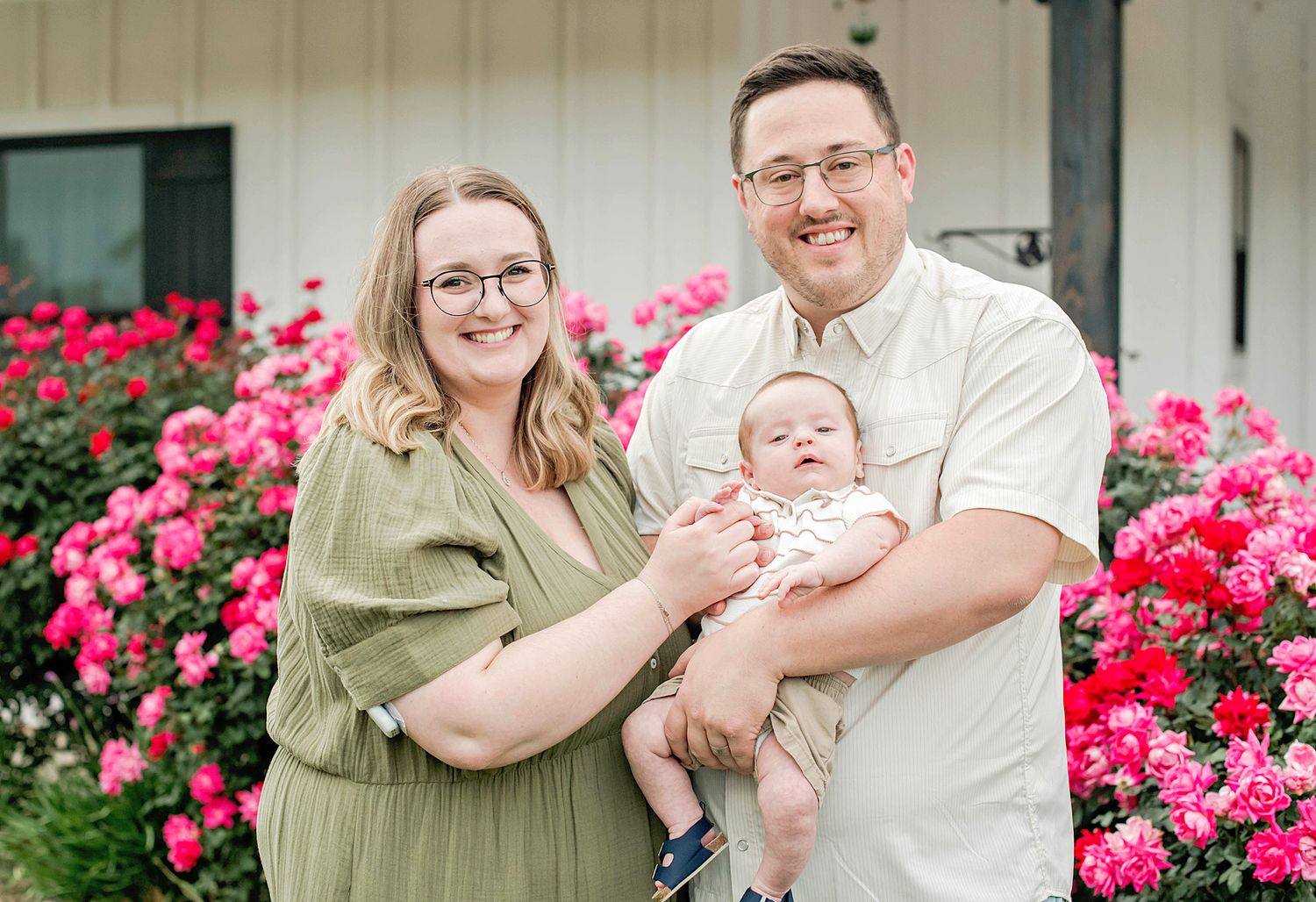 A smiling couple stands outdoors holding a newborn baby in front of vibrant pink rose bushes.