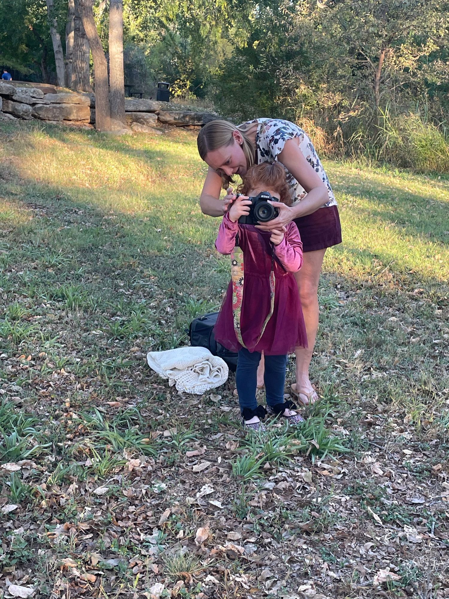 Woman teaching young girl to use a DSLR camera outdoors in a grassy park setting with trees in background.