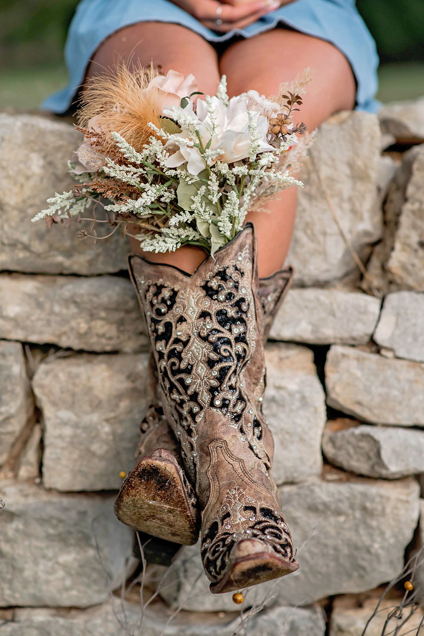 Decorative cowboy boots with floral embroidery used as a vase holding a wildflower bouquet on a stone wall.