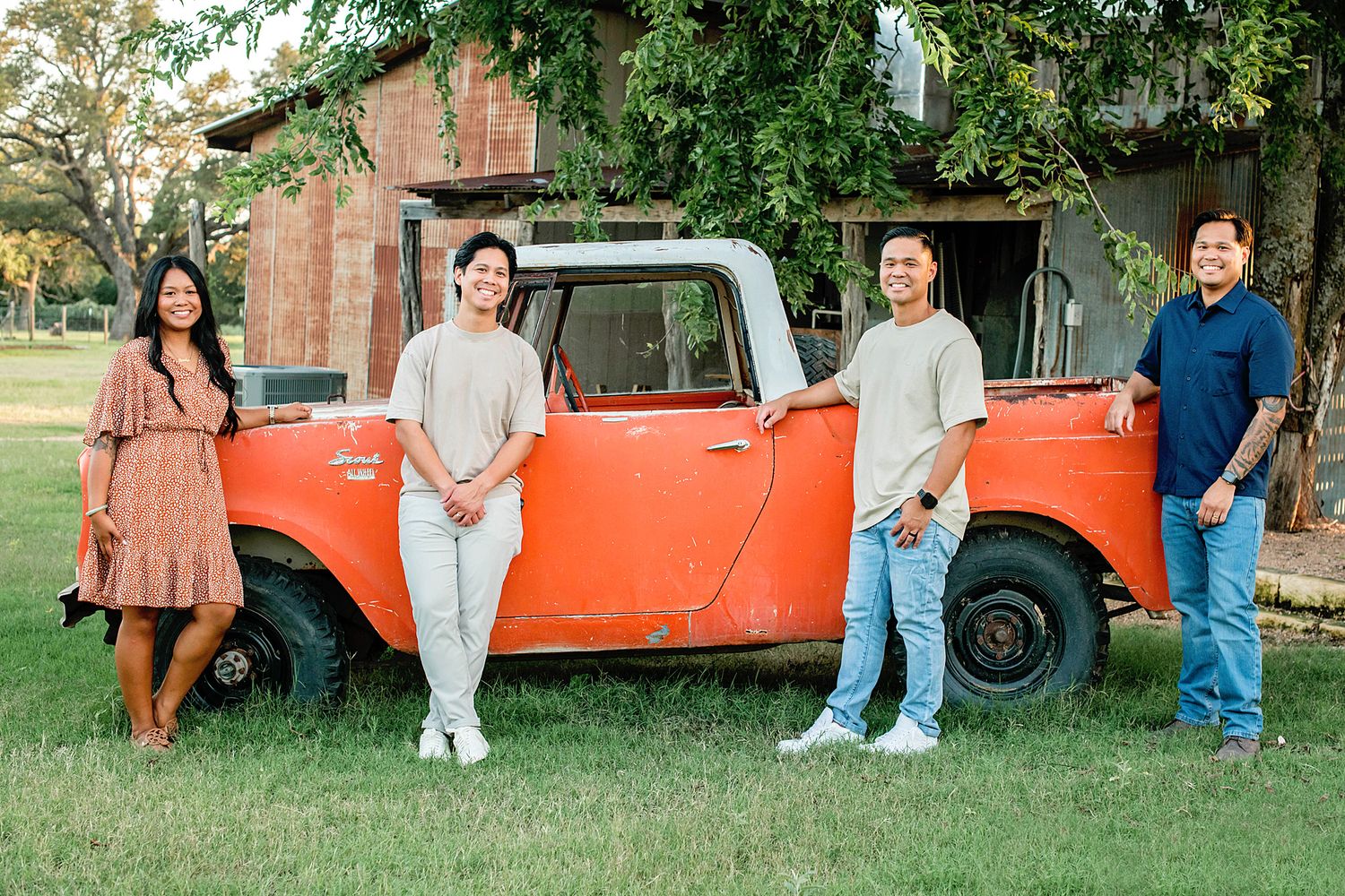 Four people smiling and posing beside a vintage orange pickup truck on a green lawn near a rustic barn.
