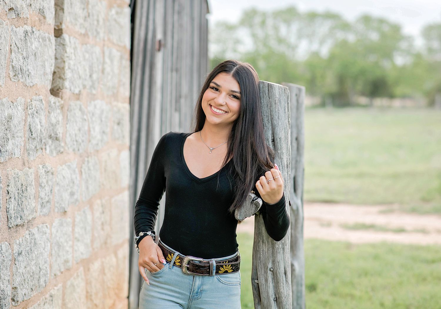Young woman in black top and jeans smiling outdoors near rustic barn and wooden fence on a farm.