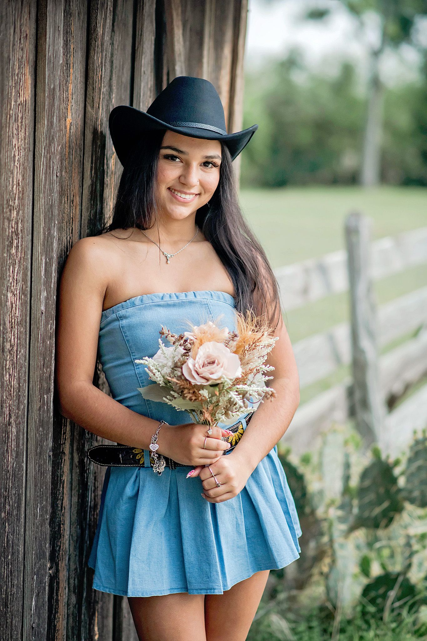 Young woman in blue strapless dress and black cowboy hat holding a wildflower bouquet, leaning against a rustic barn.