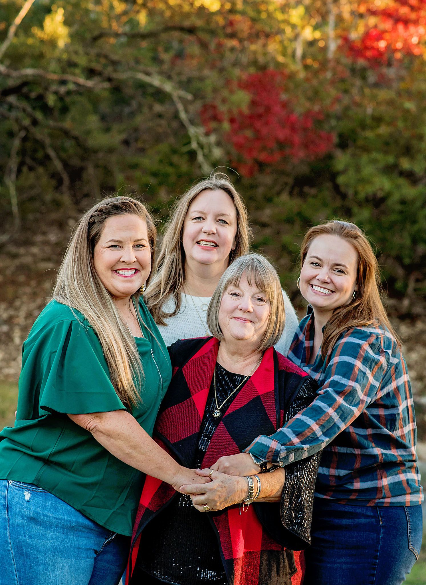 Four women smiling together outdoors in fall setting, holding hands in warm family portrait.