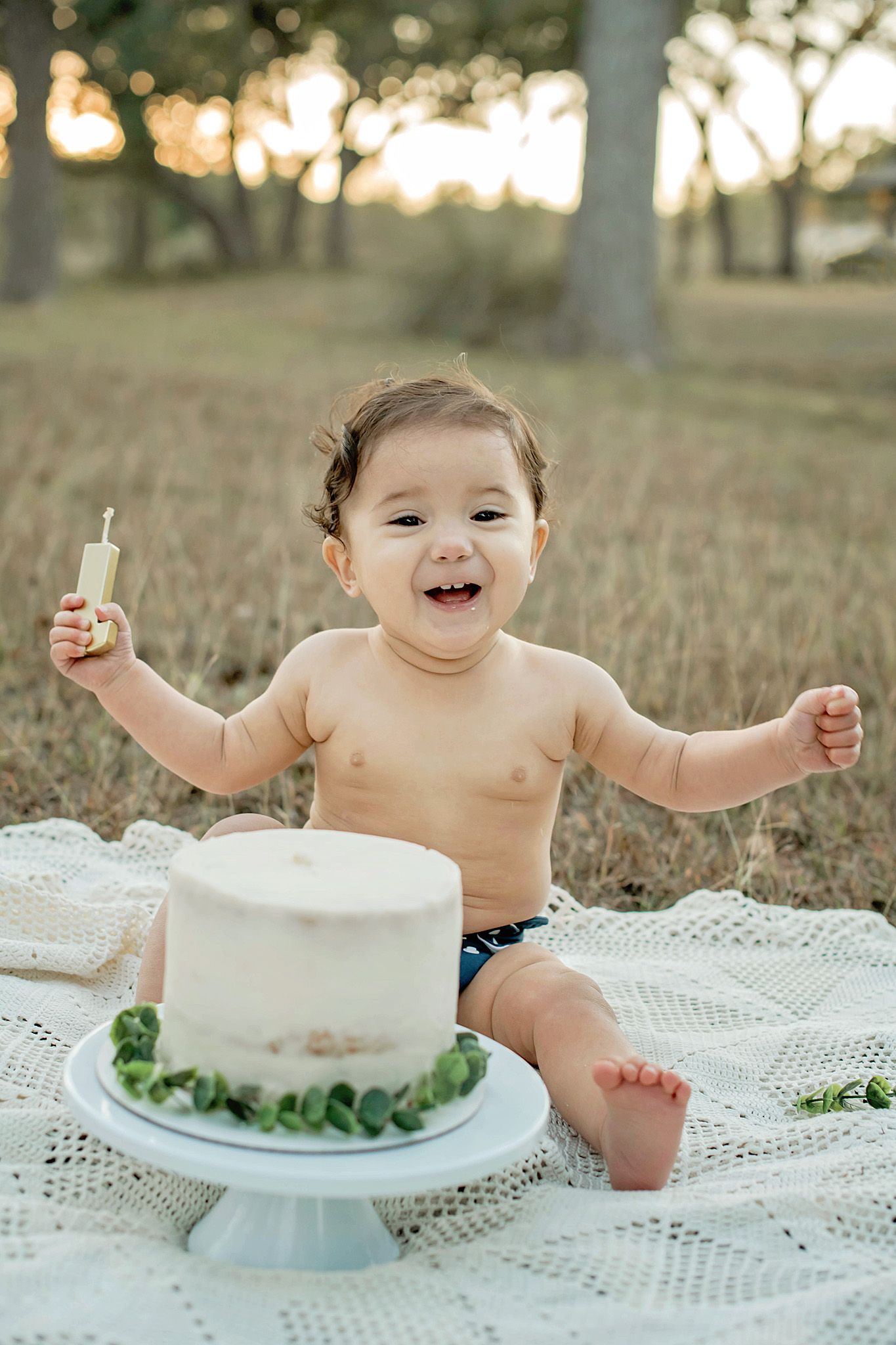 Baby smiling at first birthday cake smash outdoors on white blanket, holding candle, white cake on stand.