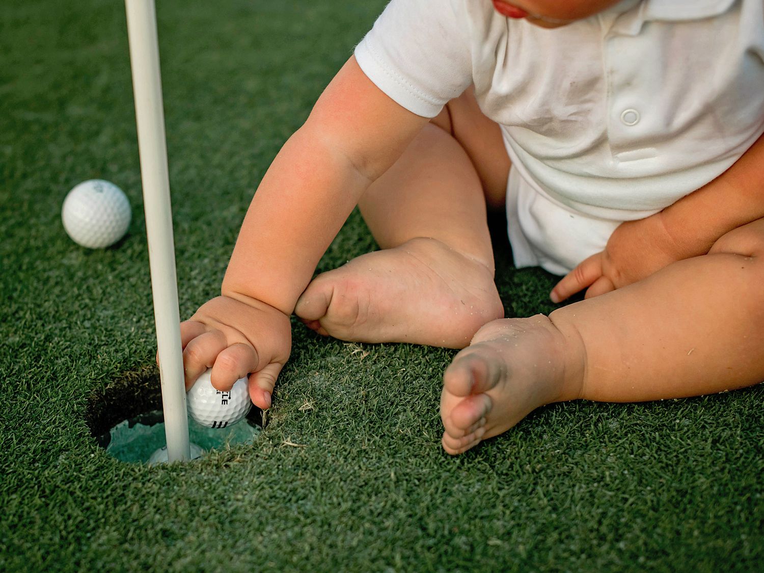 Close-up view of small child playing mini golf on artificial turf, placing ball near hole with white flag marker.
